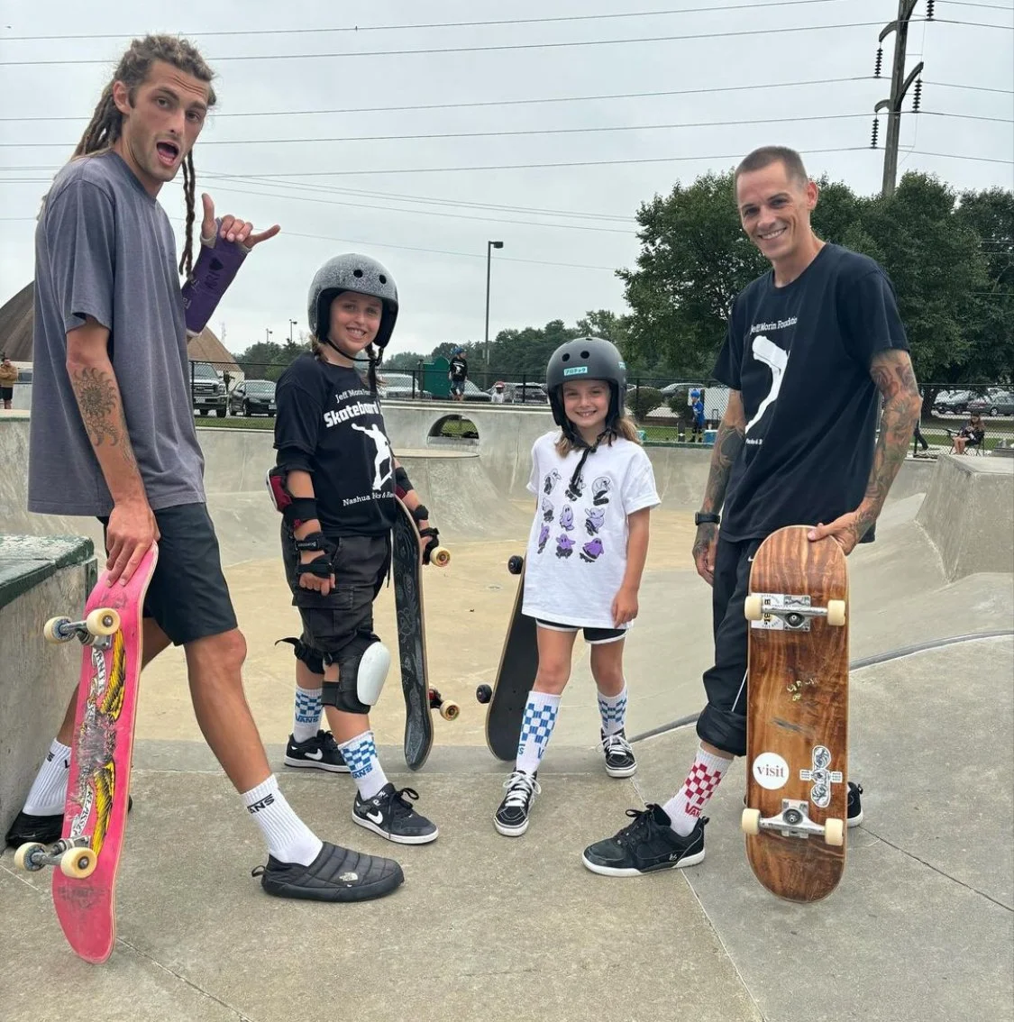 Four people with skateboards at a skatepark, two children wearing helmets and gear, two adults holding skateboards, all smiling.