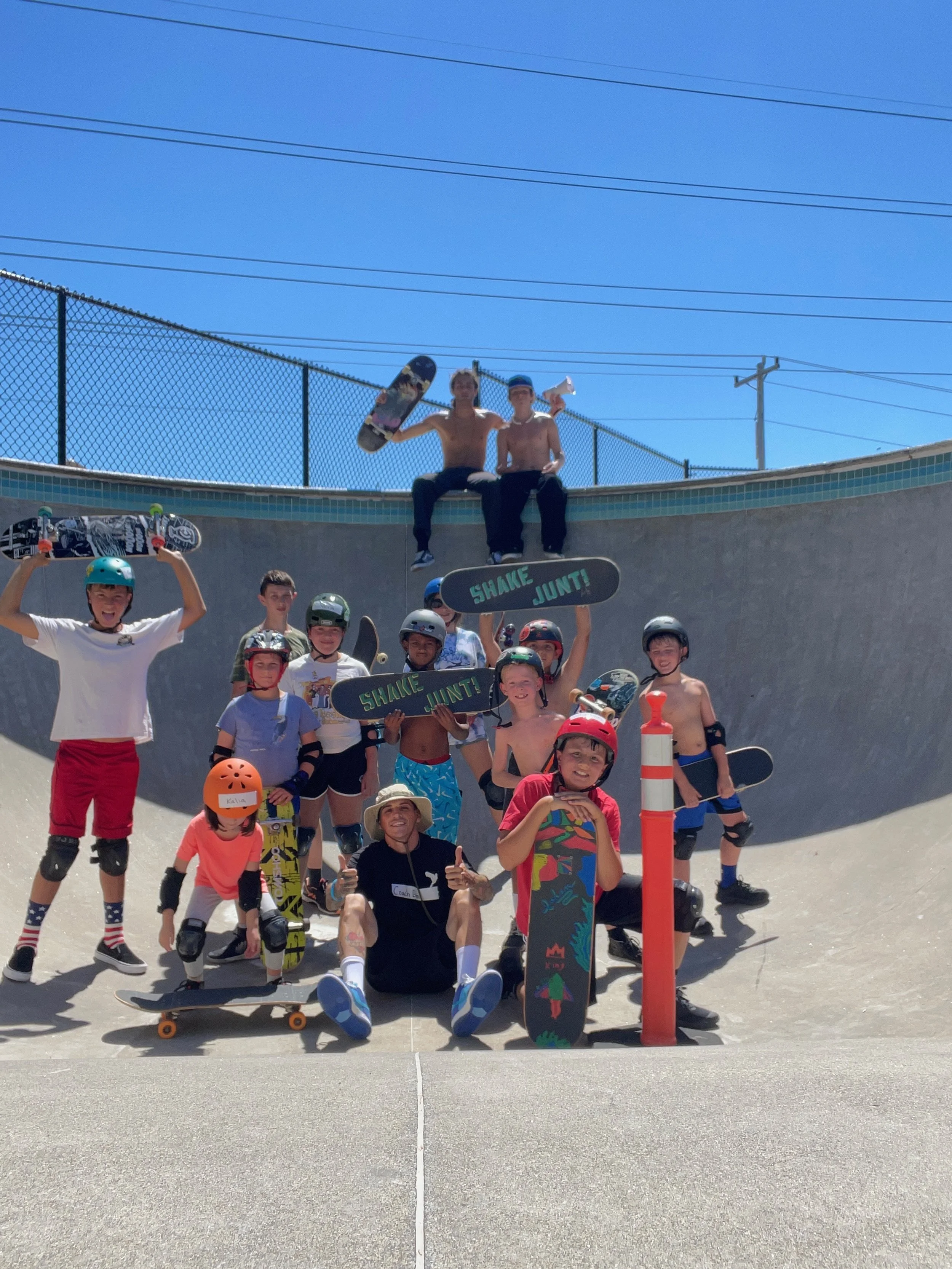 Group of kids and teenagers at a skate park, some holding skateboards, wearing helmets and protective gear, with a fence and power lines in the background on a sunny day.