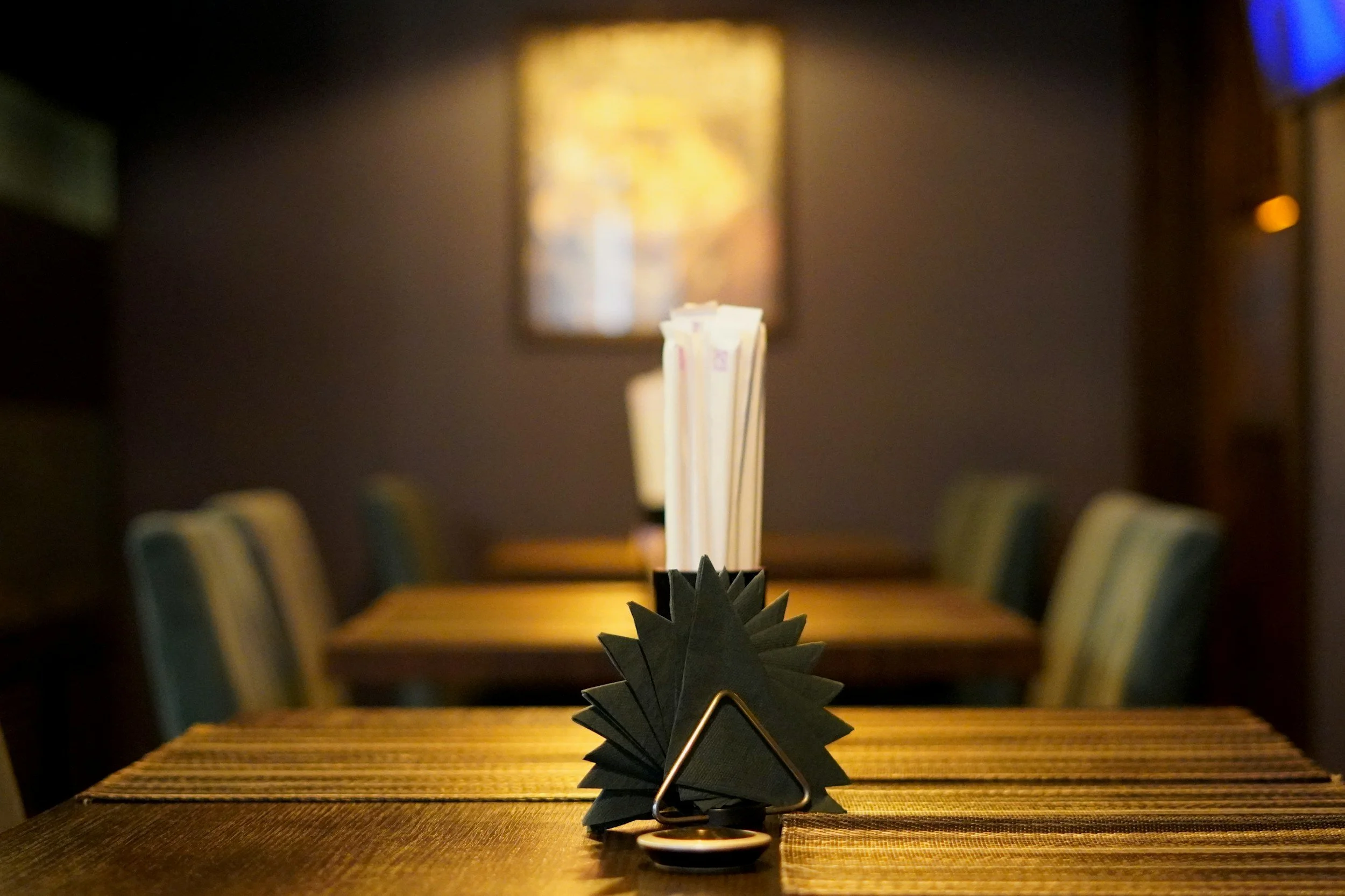 A black napkin holder with napkins and straws on a wooden table in a restaurant with chairs in the background.