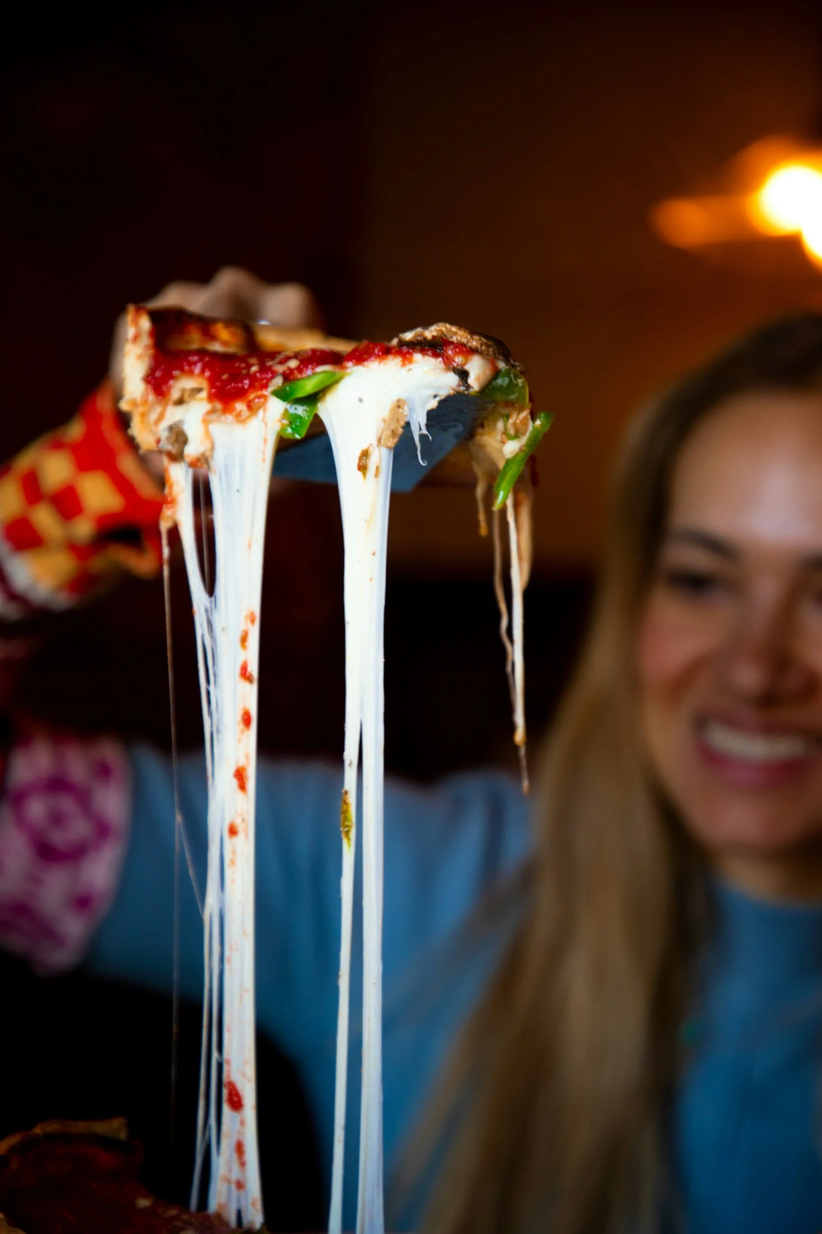 Person holding up a slice of pizza with melted cheese stretching from the slice, in focus, with a smiling woman in the background.