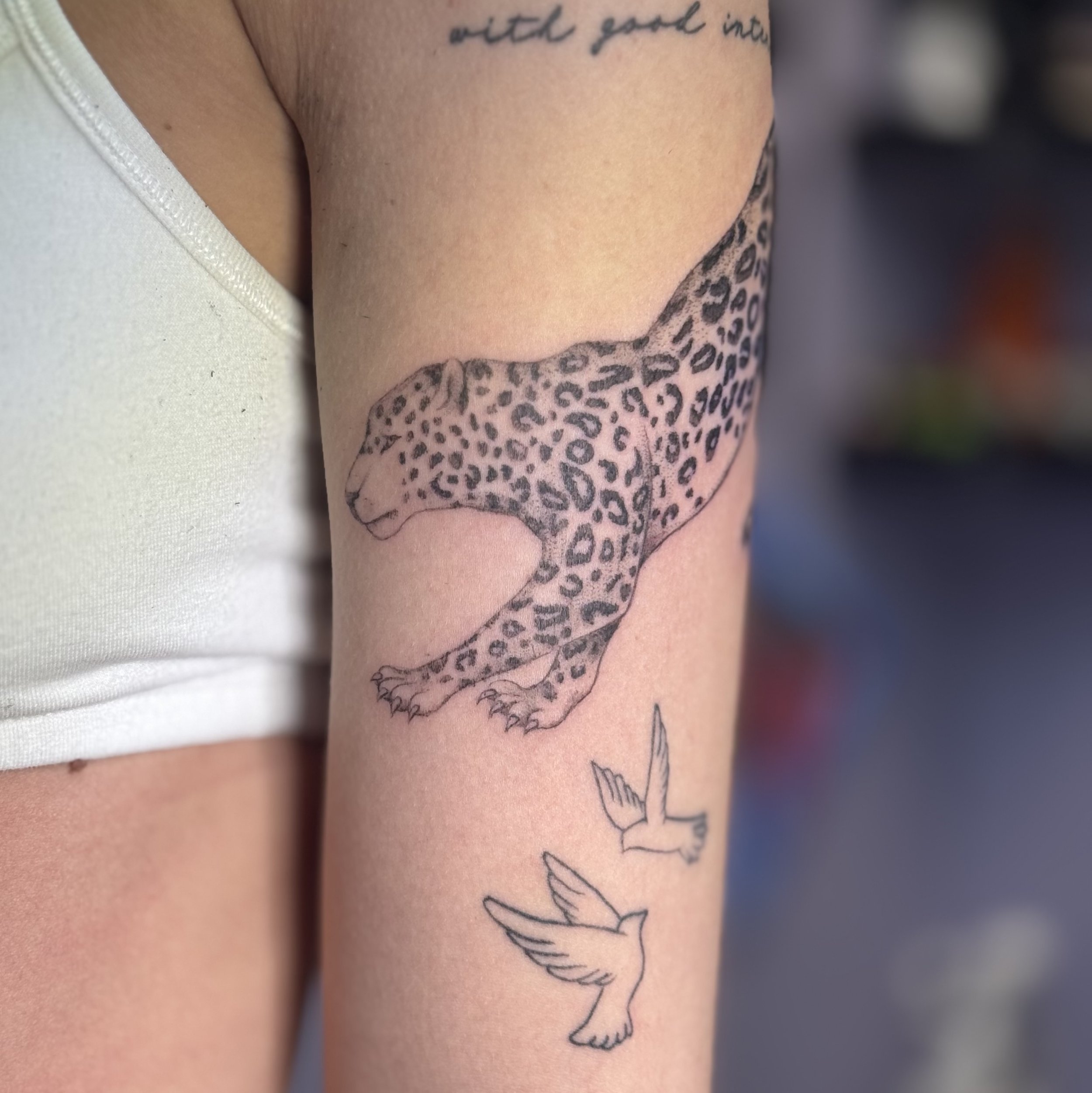 Tattoo of a leaping jaguar with leopard spots on a person's upper arm, accompanied by two outline hummingbirds below it.