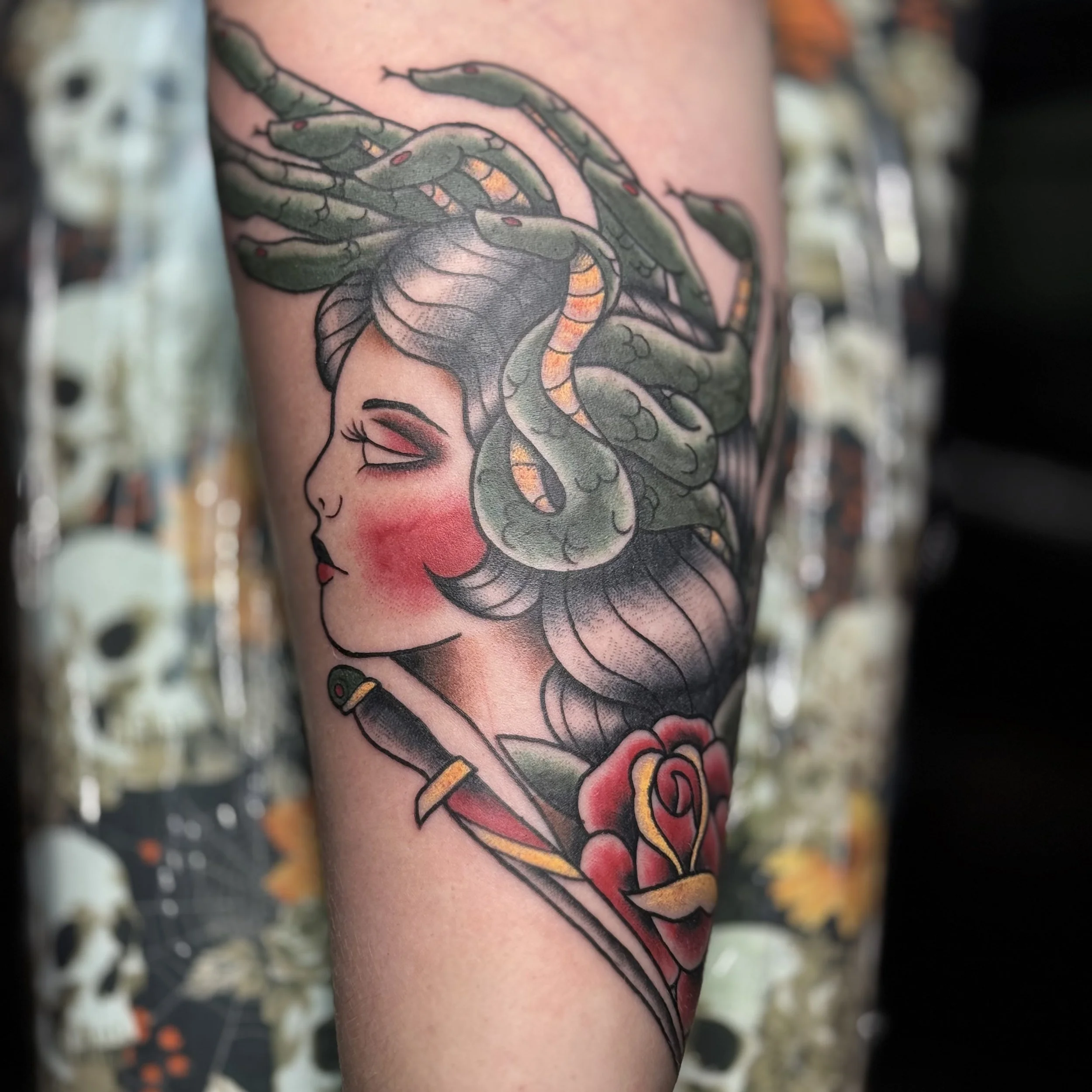 Tattoo of a woman's face with closed eyes, red blush on cheeks, dark hair, and a snake slithering across her head, with a rose flower and a dagger.