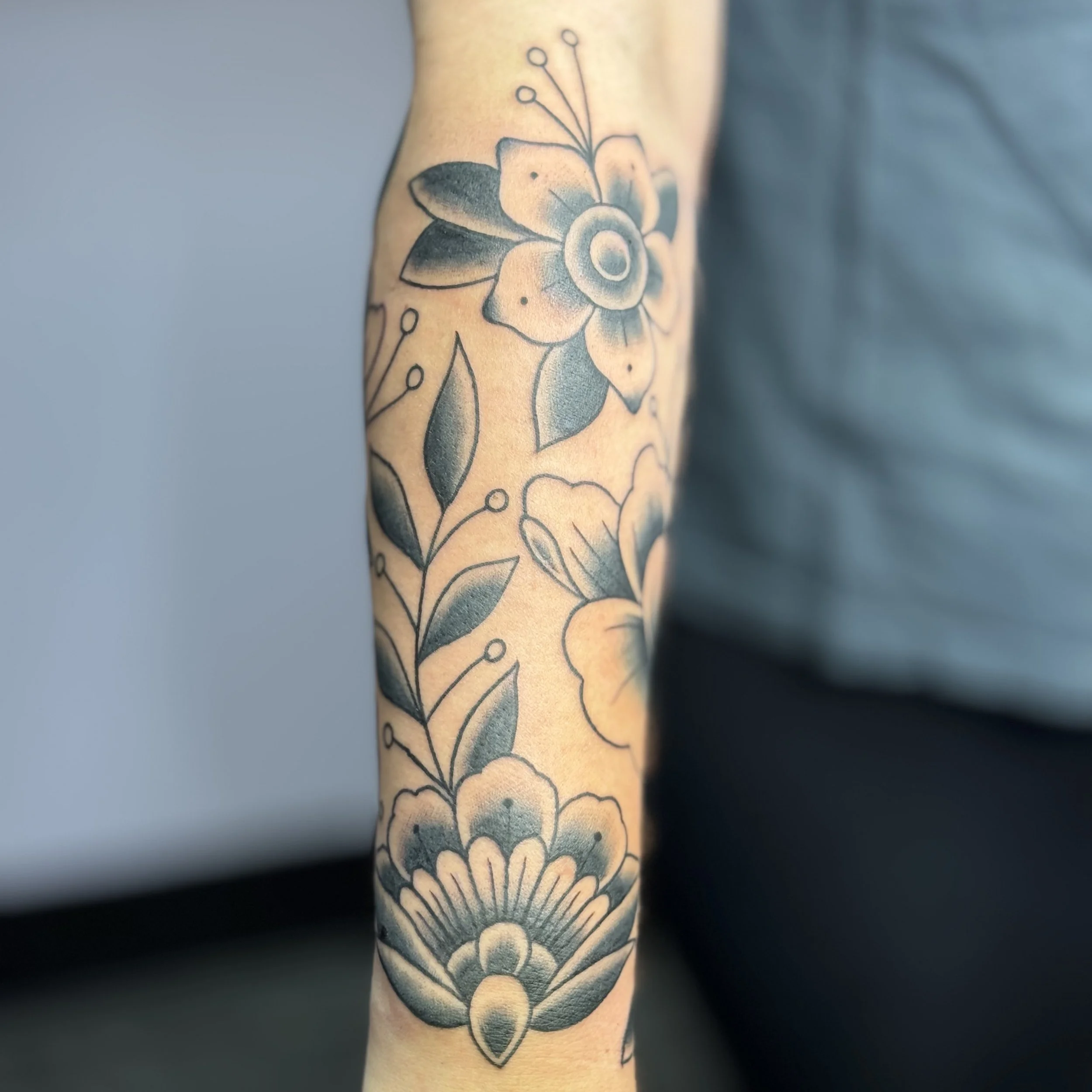 Black and gray floral tattoo on an arm, featuring a large flower at the top, leaves, and other floral patterns extending down the arm.
