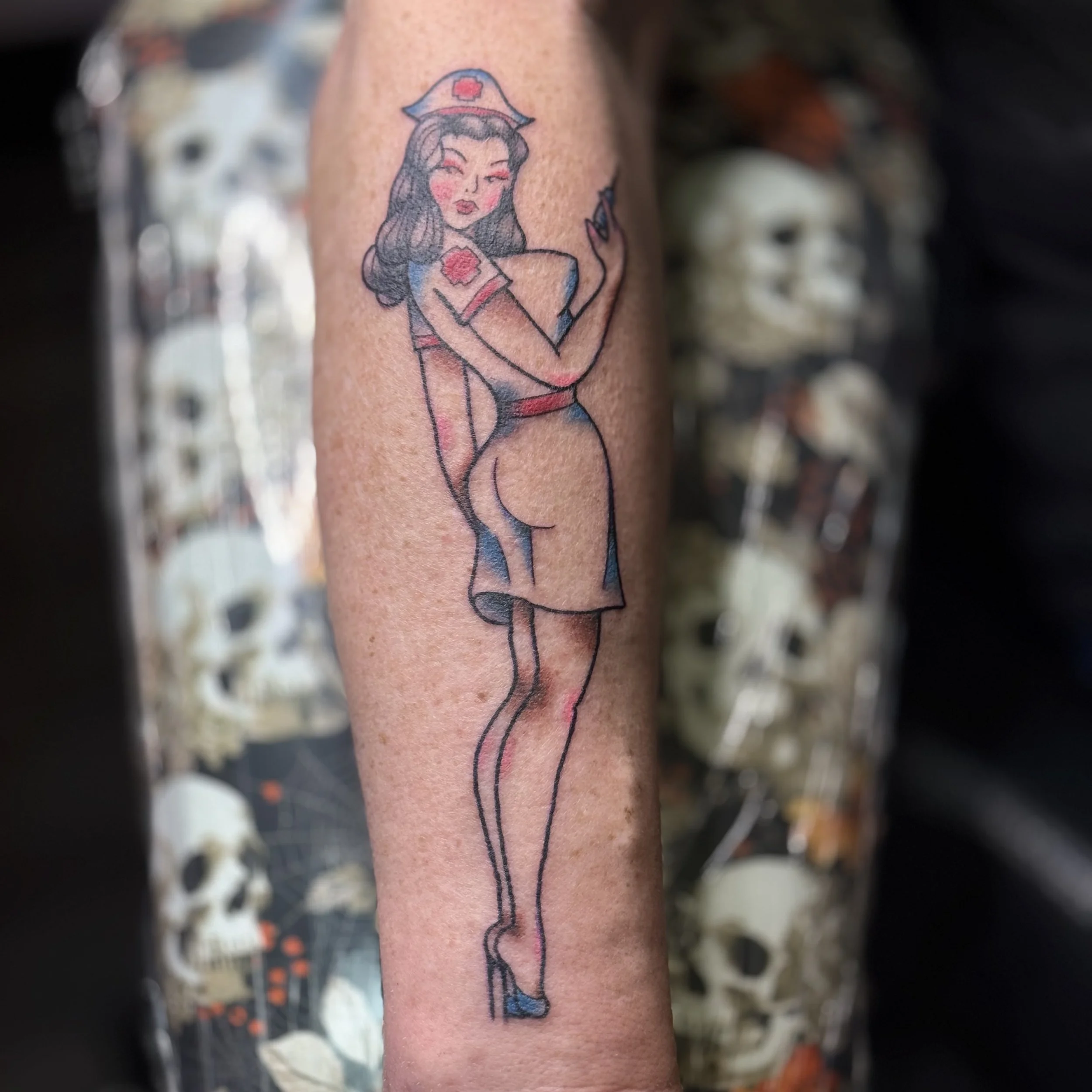 Tattoo of a woman in pin-up style nurse uniform, holding a cigarette, with dark hair and heels, on person's forearm.