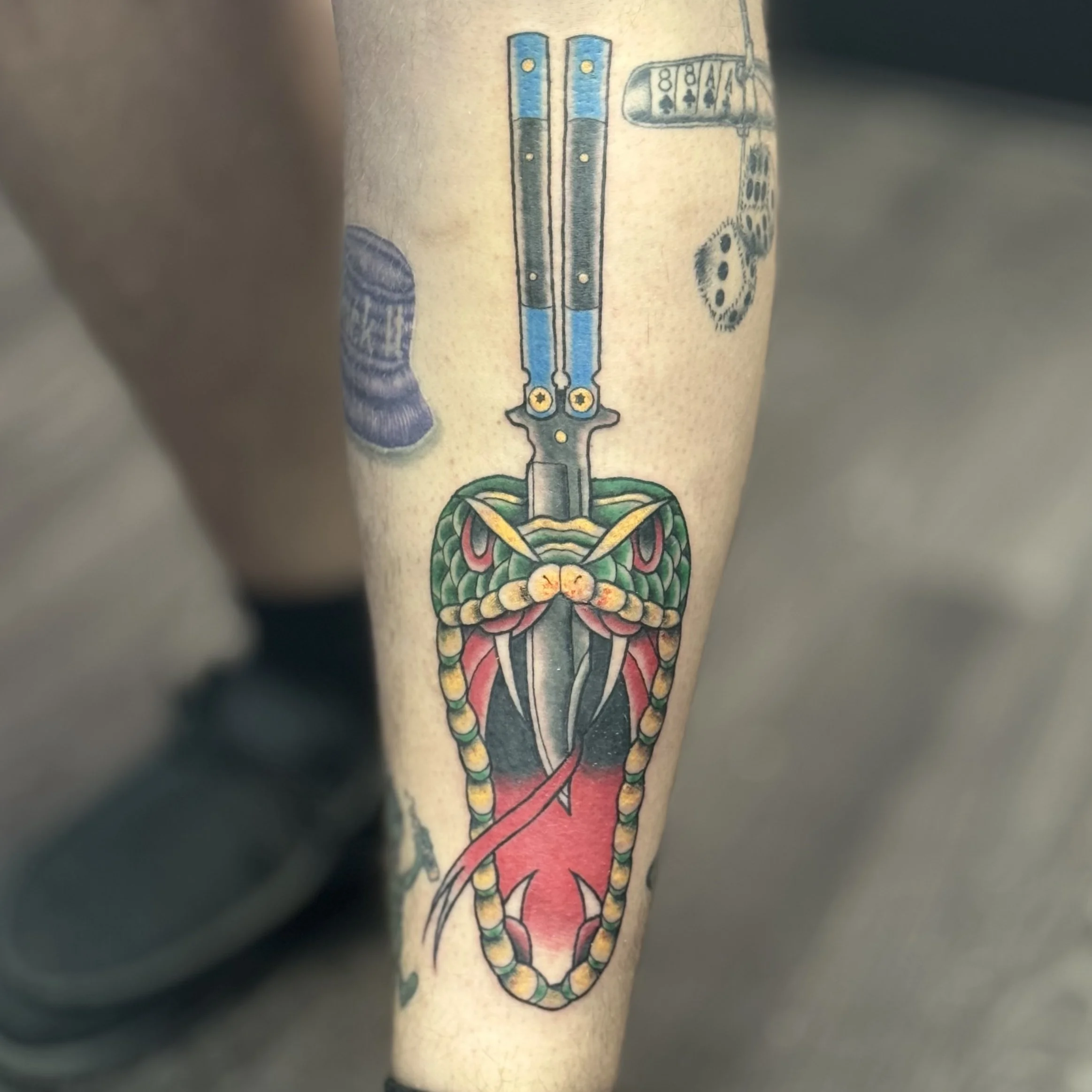 Tattoo of a tactical knife with a snake coiled around it, with the snake's head near the knife's handle and tongue extended, on a person's forearm.