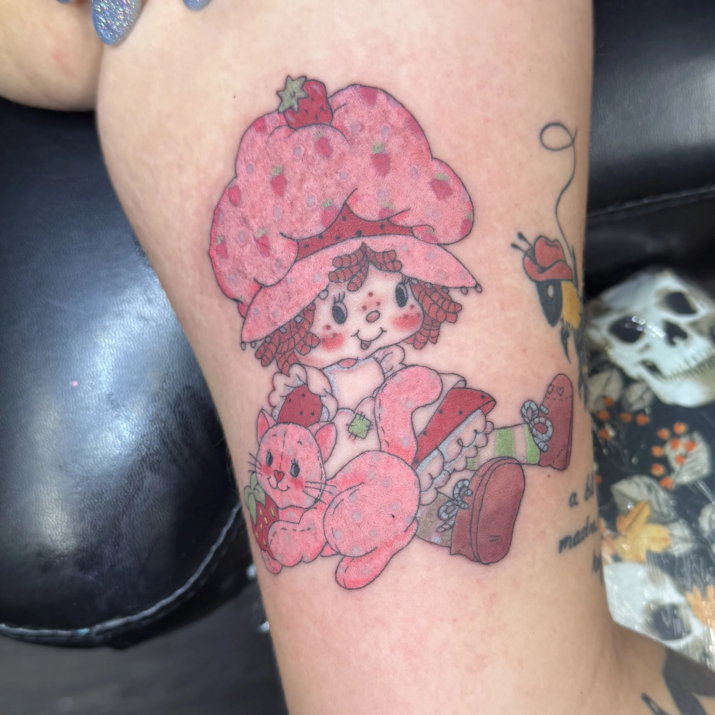 Colorful tattoo of a cute girl with strawberry bonnet, holding a pink bunny, surrounded by strawberries and a teddy bear, with some text on the lower part.
