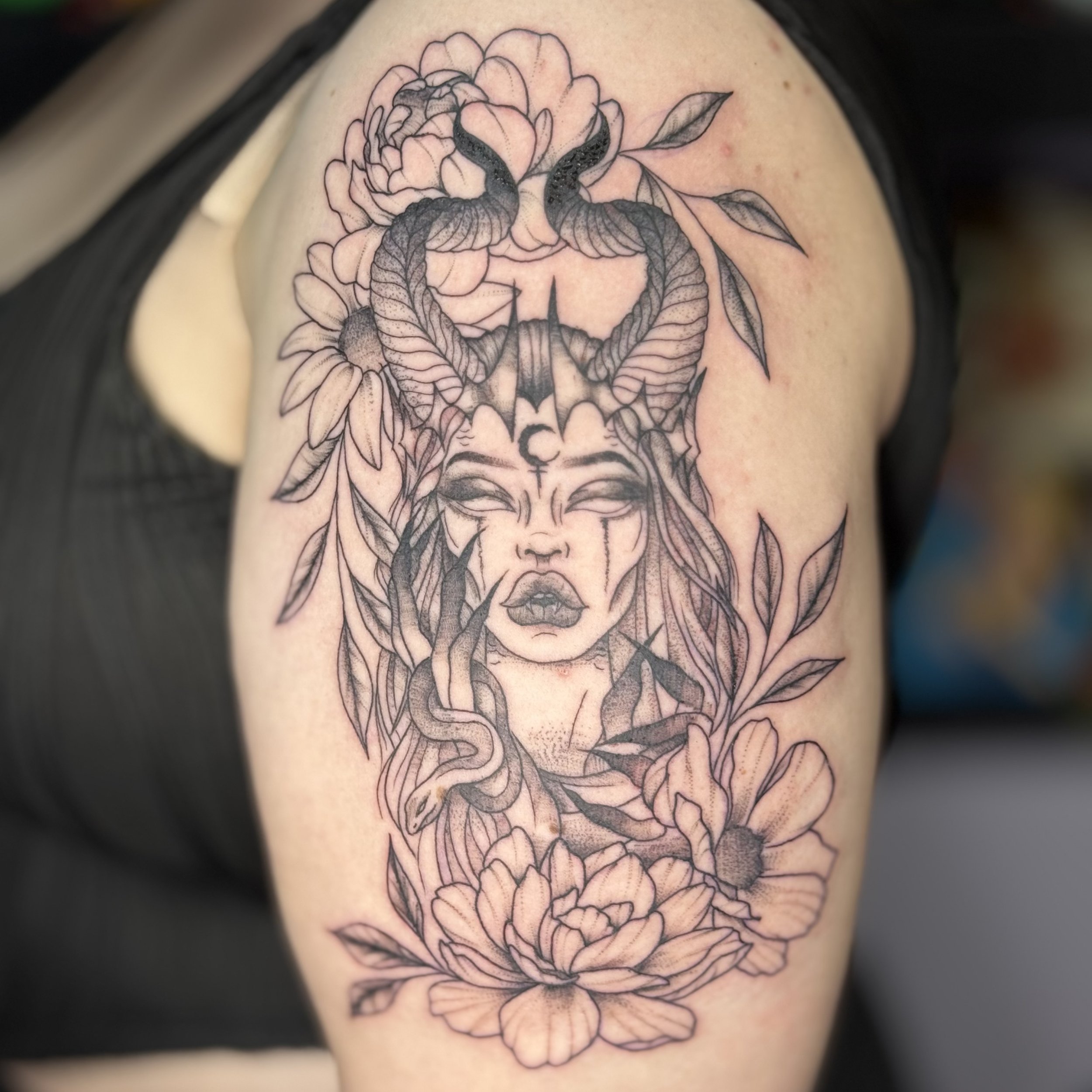 Black and gray tattoo on upper arm of a woman with long hair, closed eyes, and tears, surrounded by flowers, a snake, and horns.