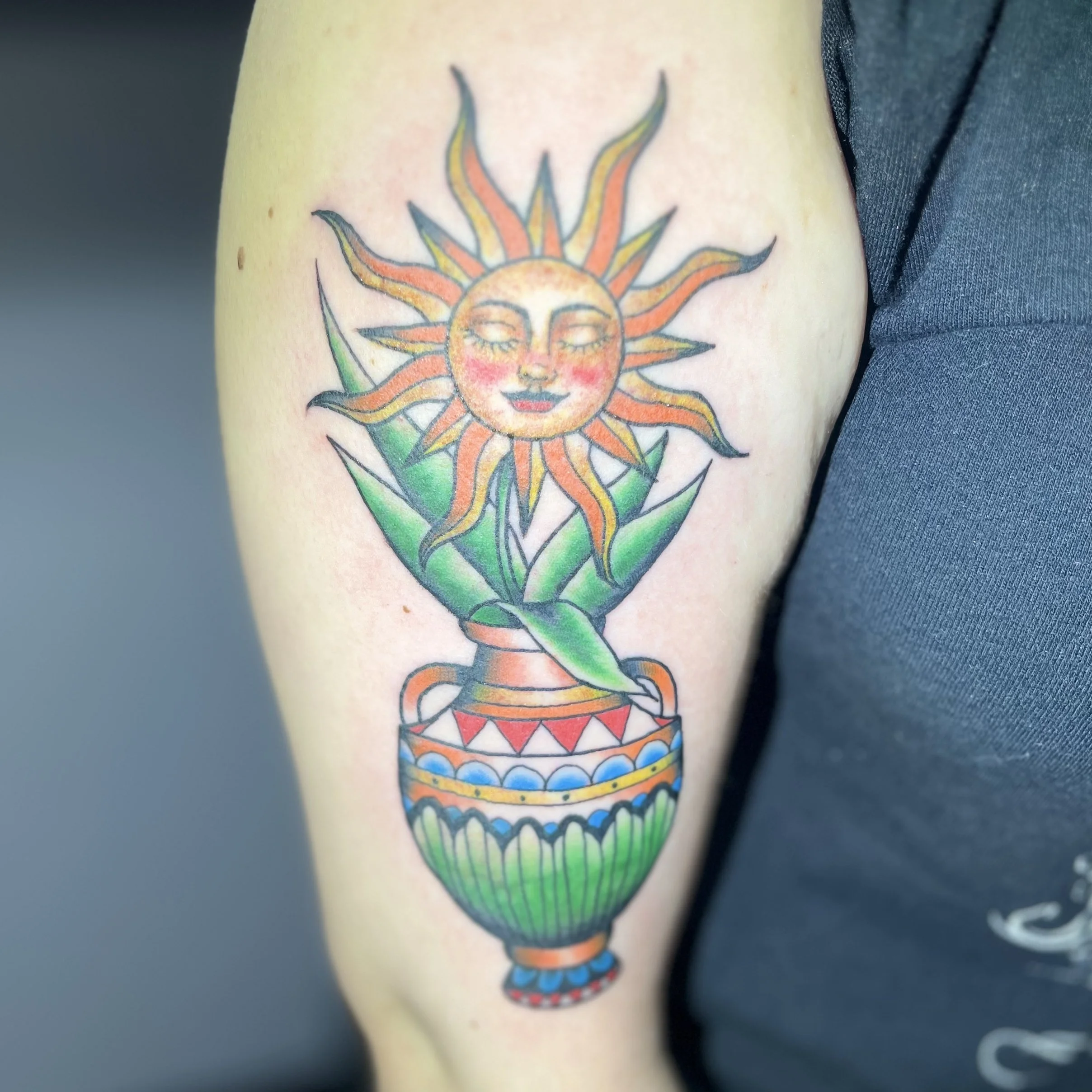 Tattoo of a smiling sun with a face, radiating orange and yellow rays, in a decorative vase with green, red, blue, and orange patterns, on a person's arm.