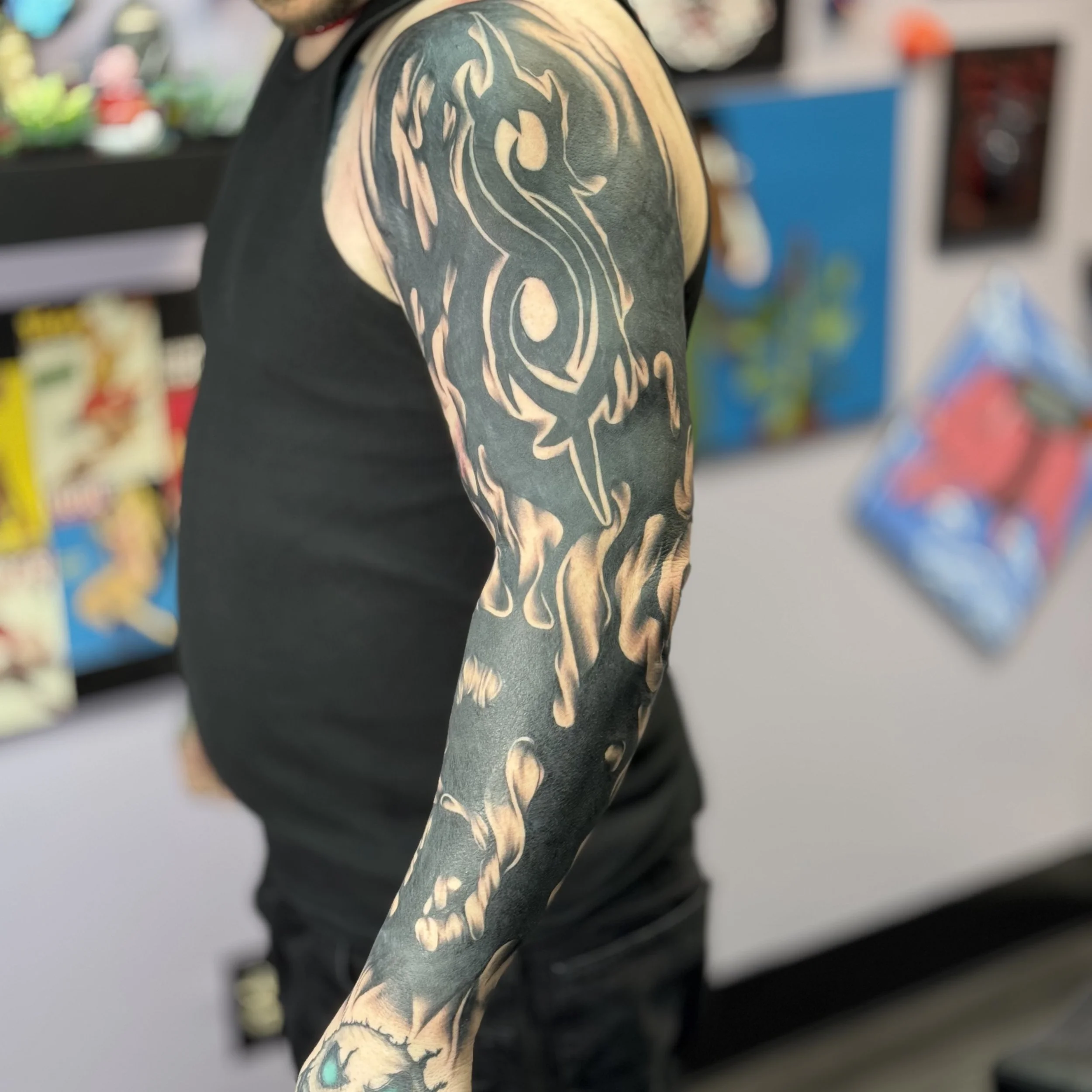 Close-up of a person's arm with a black and beige tribal-style tattoo of a dragon or wolf on the upper arm and sleeve, in a store with comic books or artwork in the background.