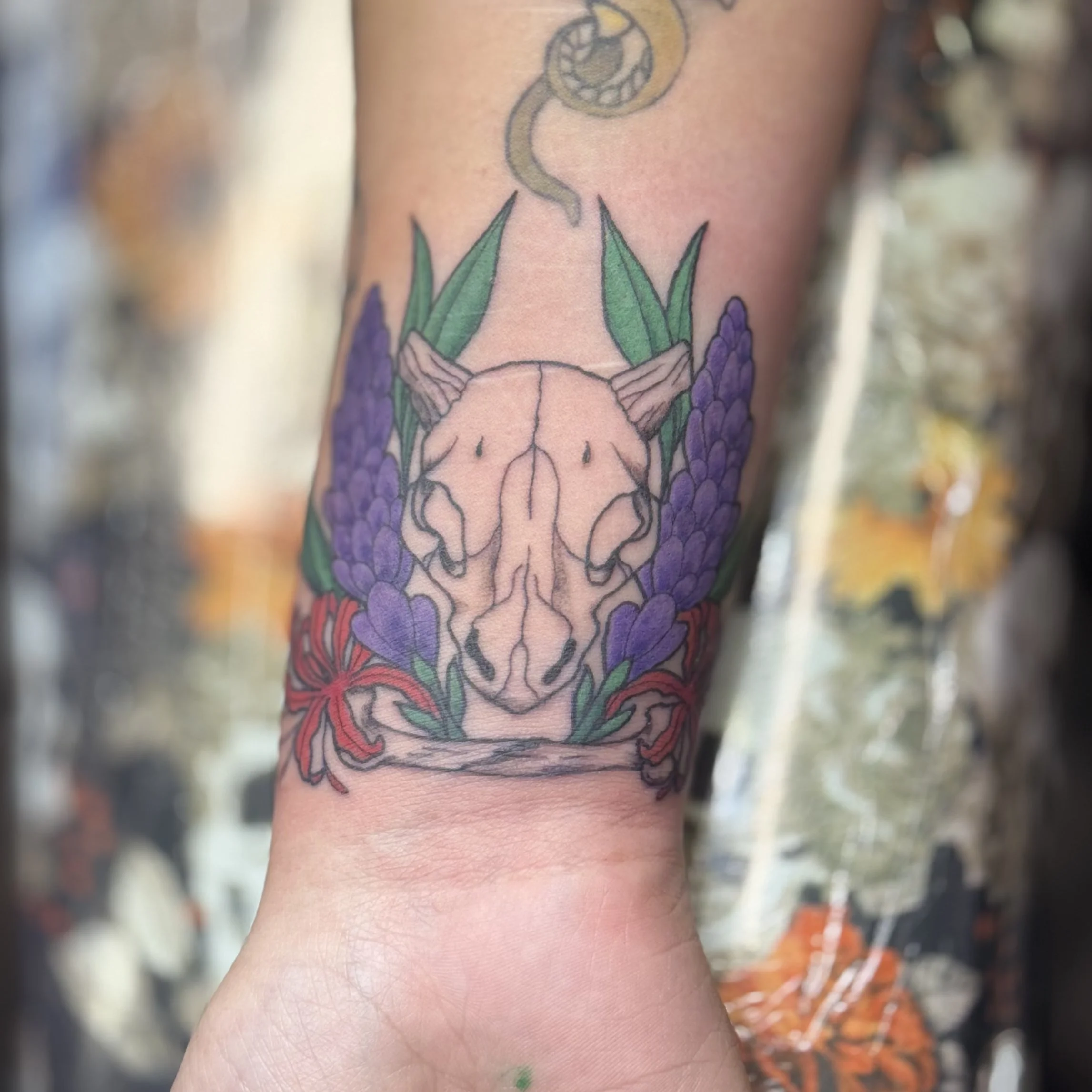 A colorful tattoo on a person's arm depicting a animal skull surrounded by purple flowers and green leaves, with a snake above it.