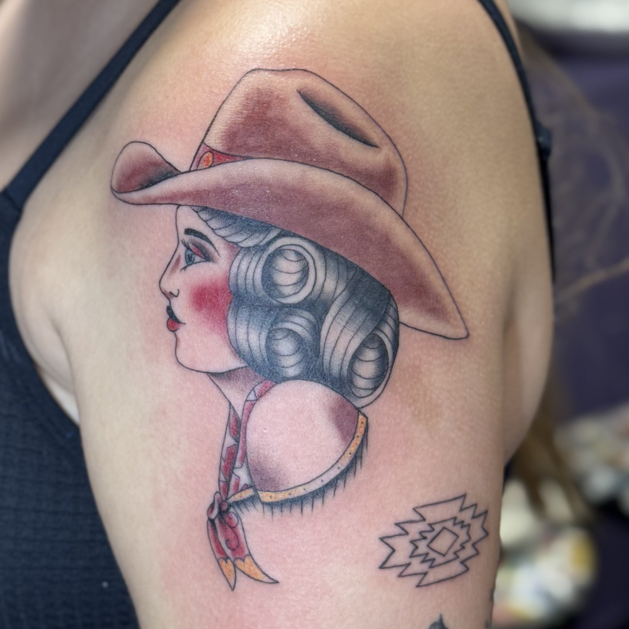 Colorful tattoo of a woman with vintage hairstyle, wearing a cowboy hat, with a red headscarf, and small decorative tattoo below.