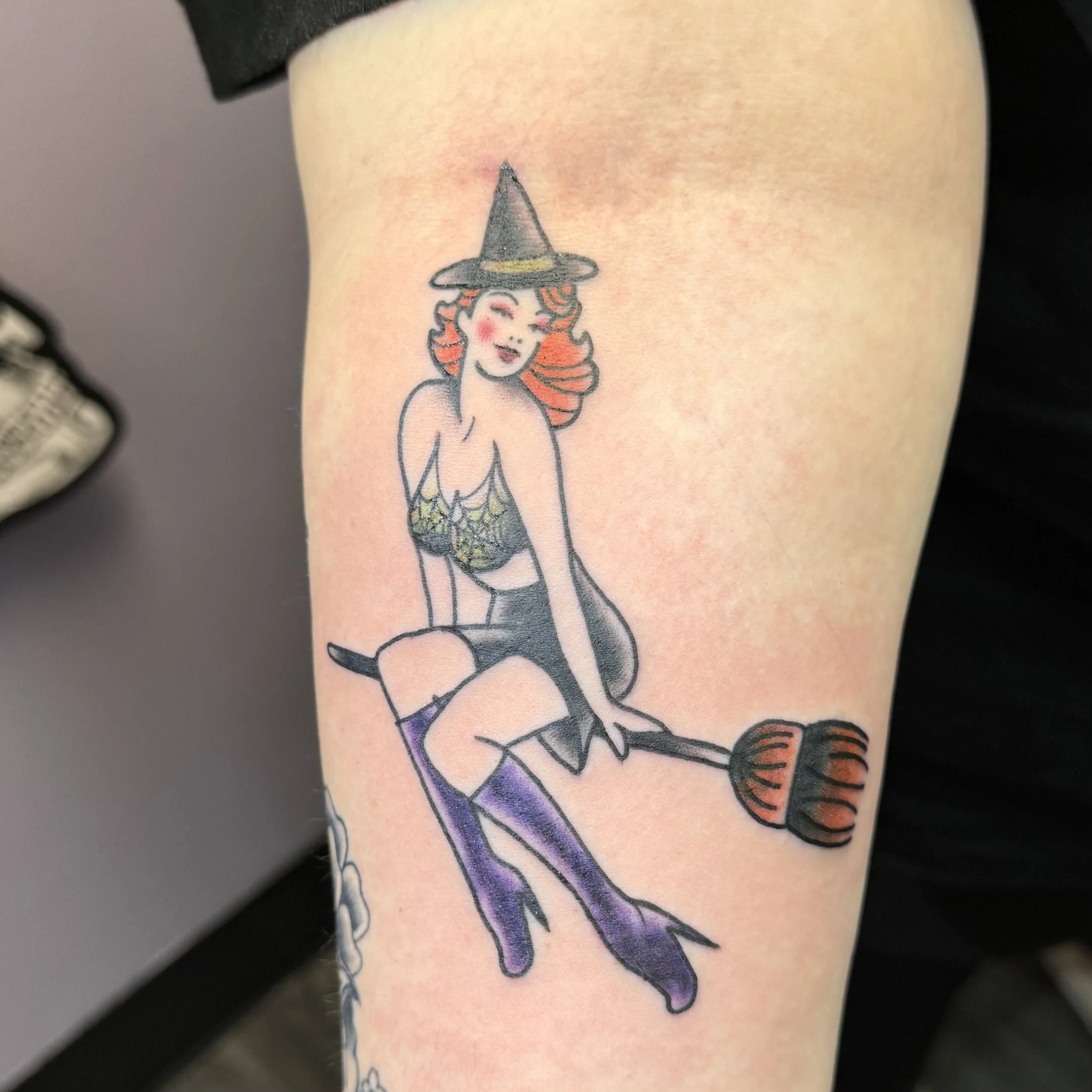 A tattoo of a red-haired witch wearing a black hat, a lace top, purple thigh-high stockings, and high heels, sitting on a broomstick.