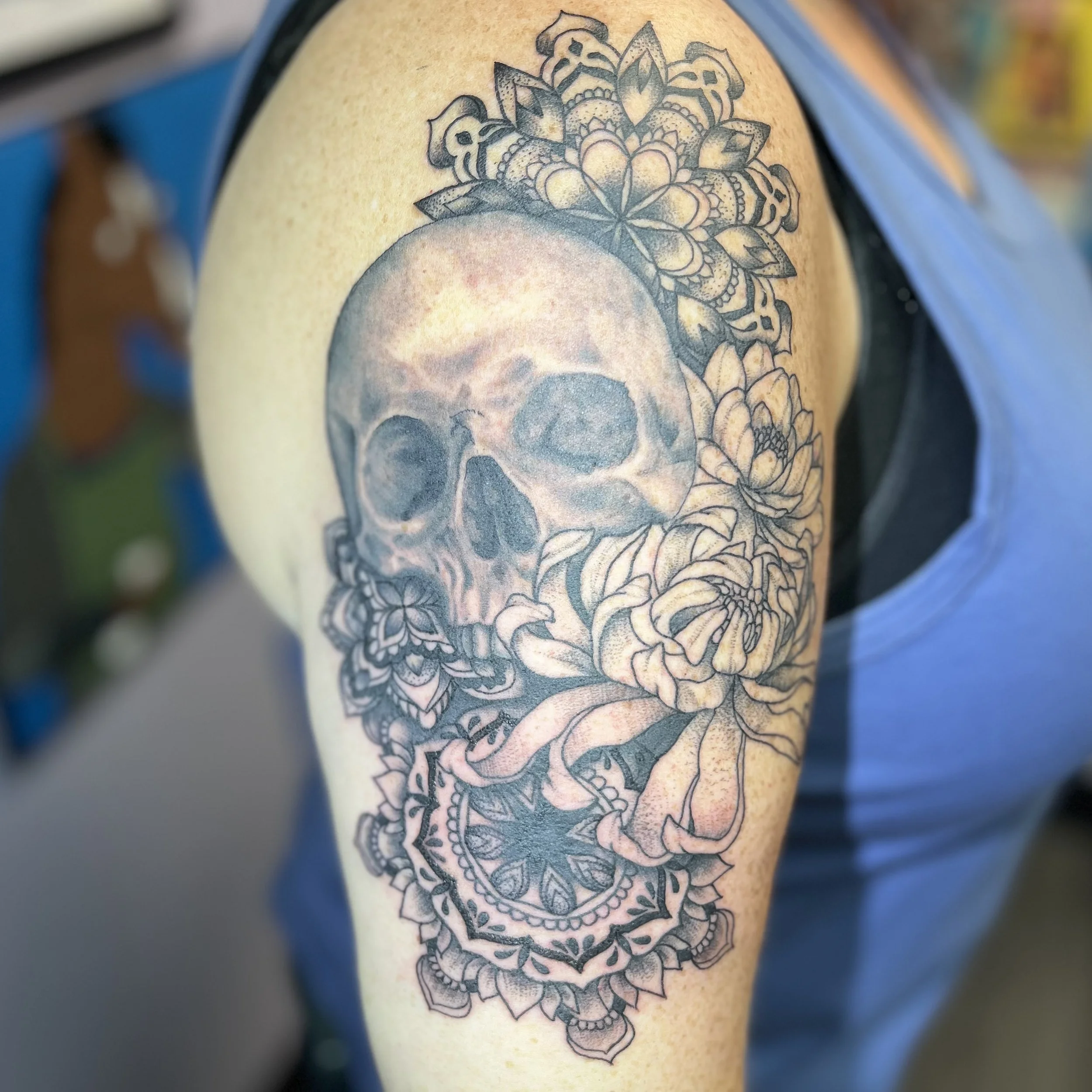 Tattoo of a skull surrounded by flowers and intricate patterns on a person's upper arm.