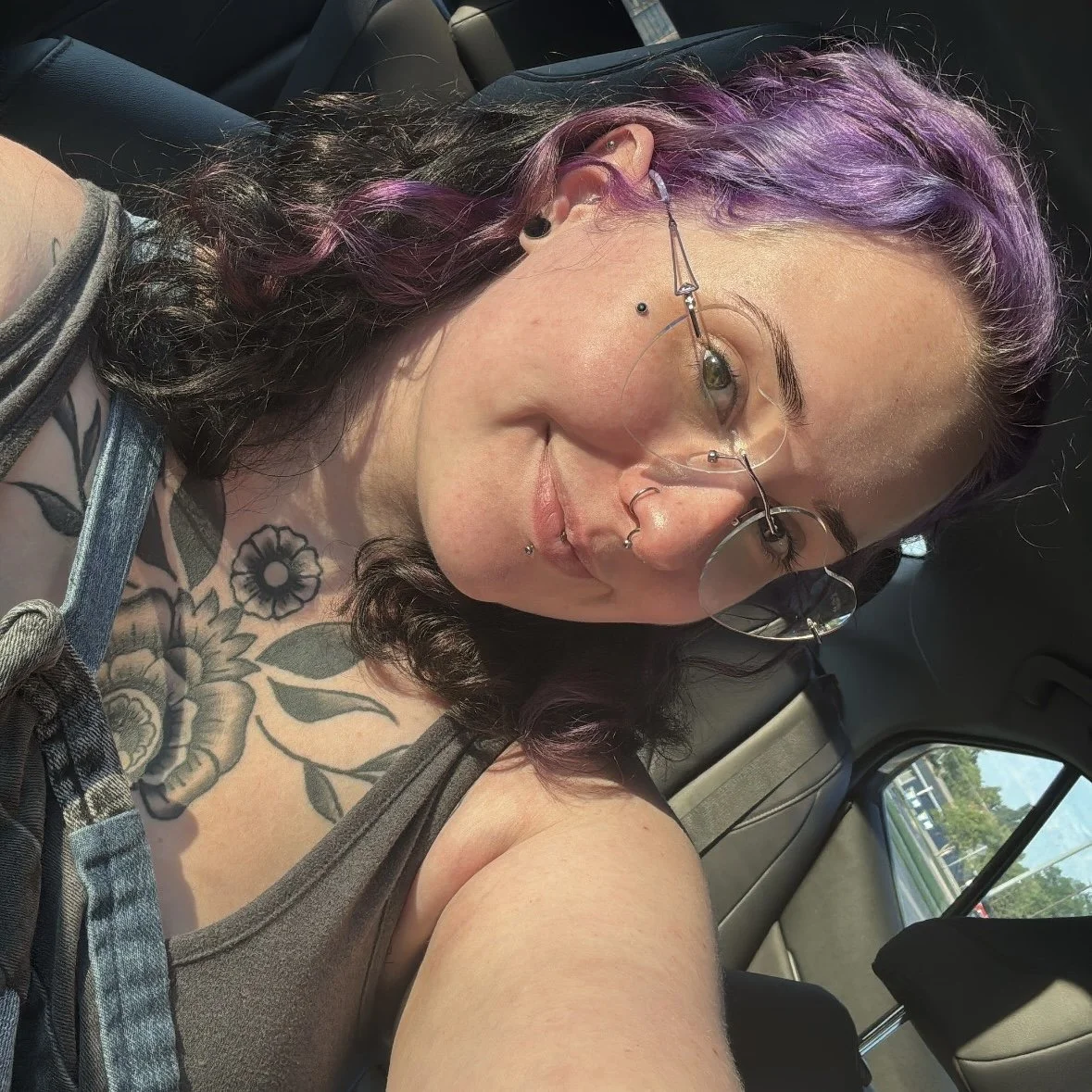 A woman with purple hair and multiple facial piercings, wearing glasses, taking a selfie in a car. She has tattoos on her shoulder and is dressed in a tank top.
