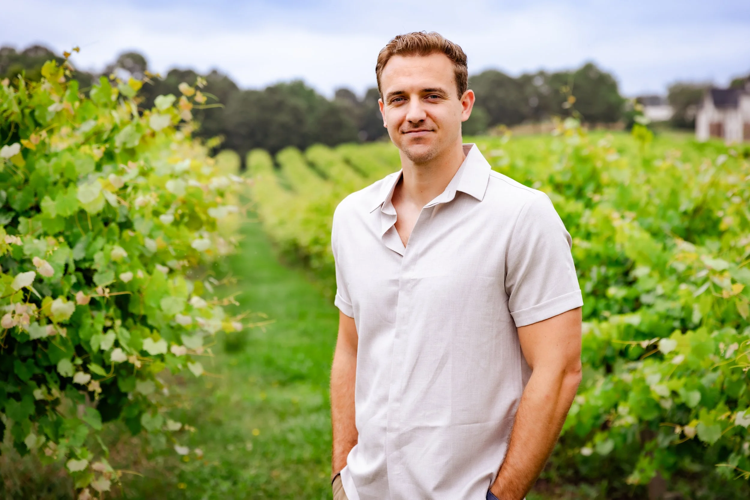 A young man in a light-colored shirt standing in a vineyard with rows of grapevines and green grass, with trees and a house in the background on a cloudy day.
