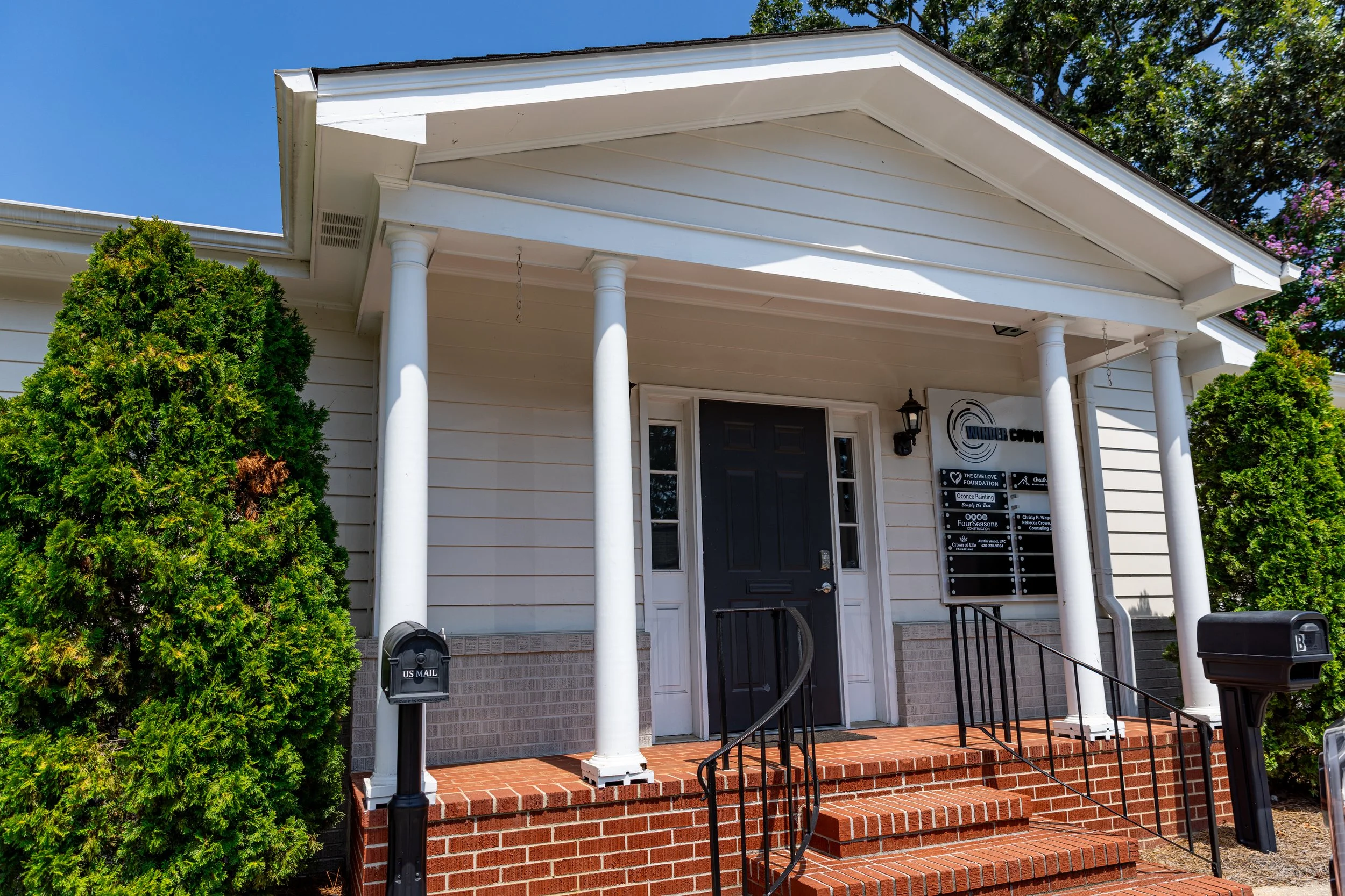 Front entrance of a white building with a black door, brick steps, columns, and surrounding greenery.