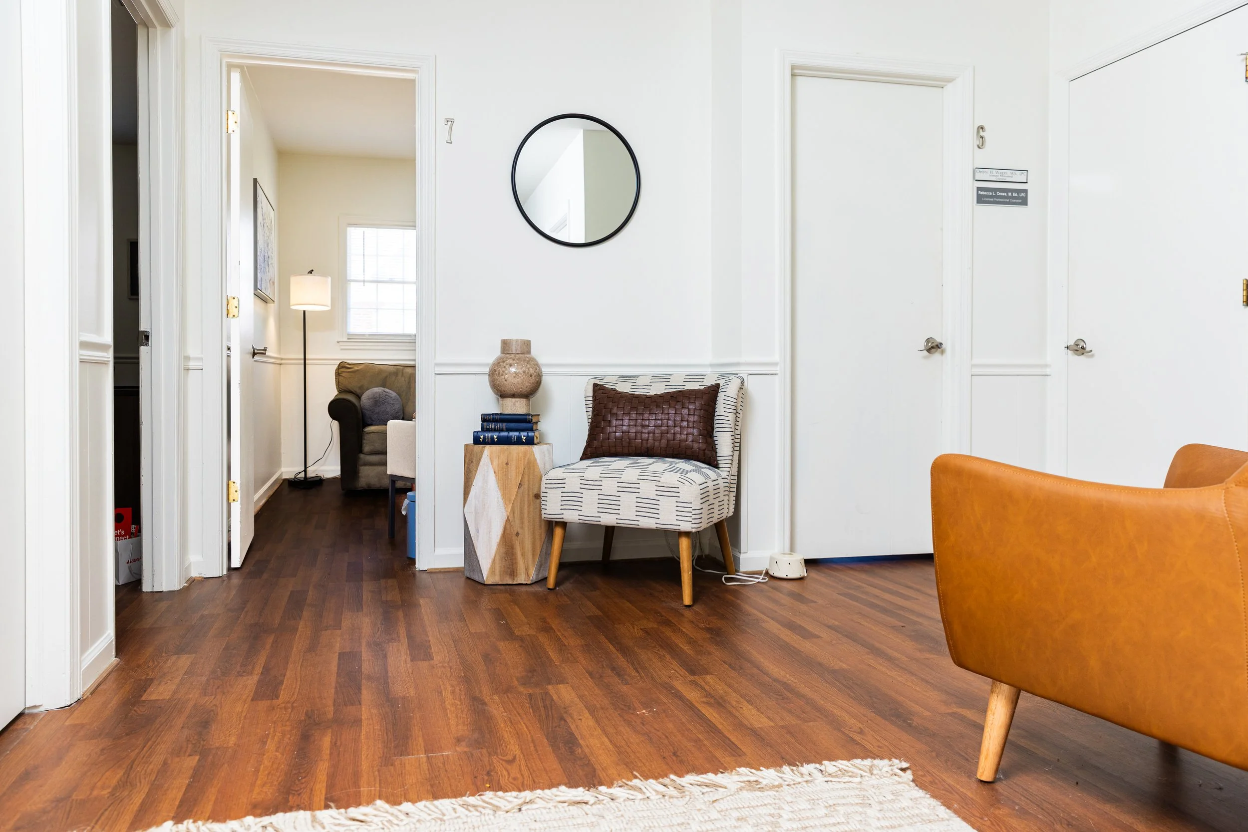 Living room with wooden floor, white walls, a patterned white chair with a brown pillow, a small wooden side table with a decorative vase, a round mirror, and a tan armchair. There is a doorway leading to a room with a brown sofa, lamp, and window.