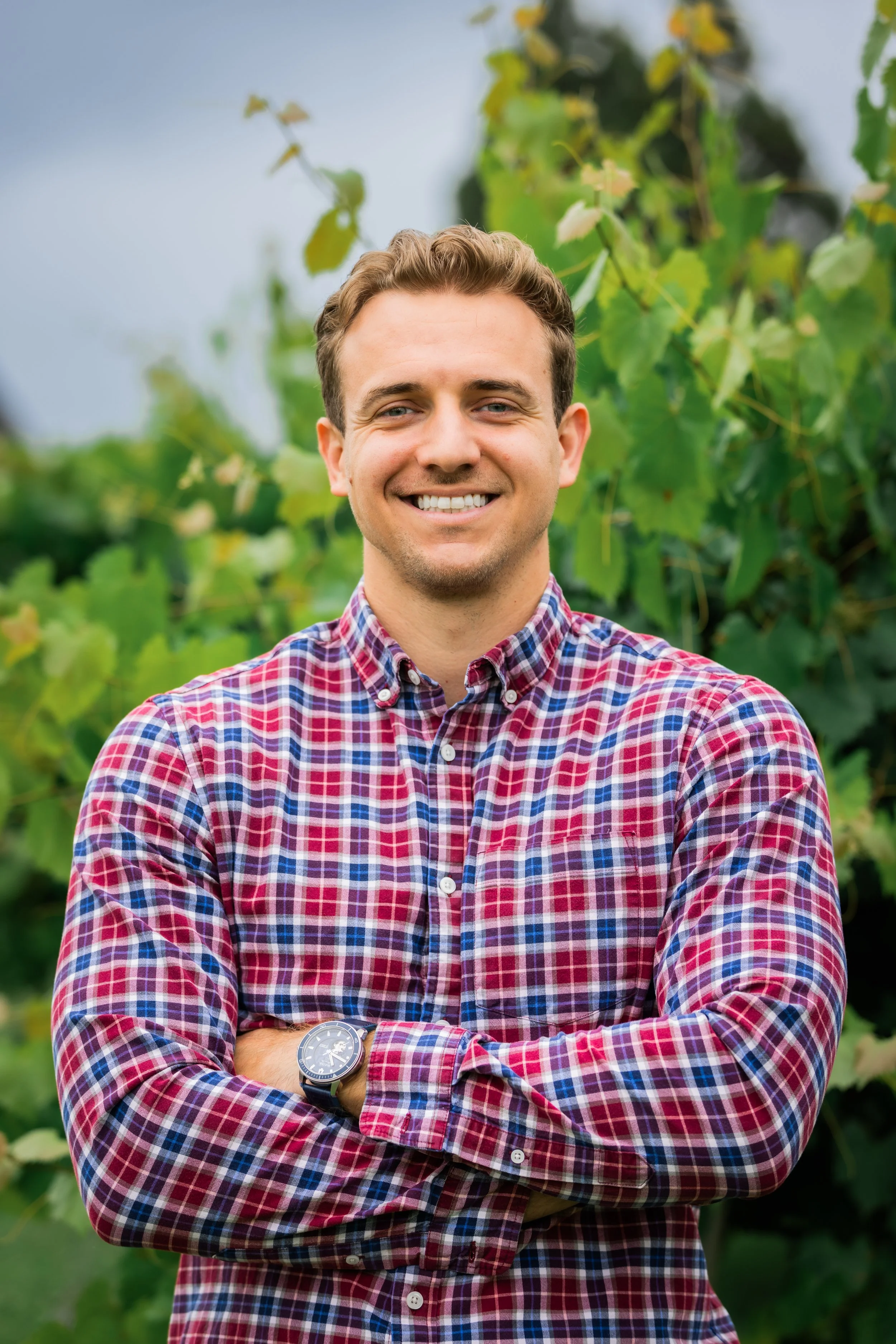 A smiling man wearing a red, blue, and white plaid shirt with his arms crossed, standing outdoors in front of green leafy plants.