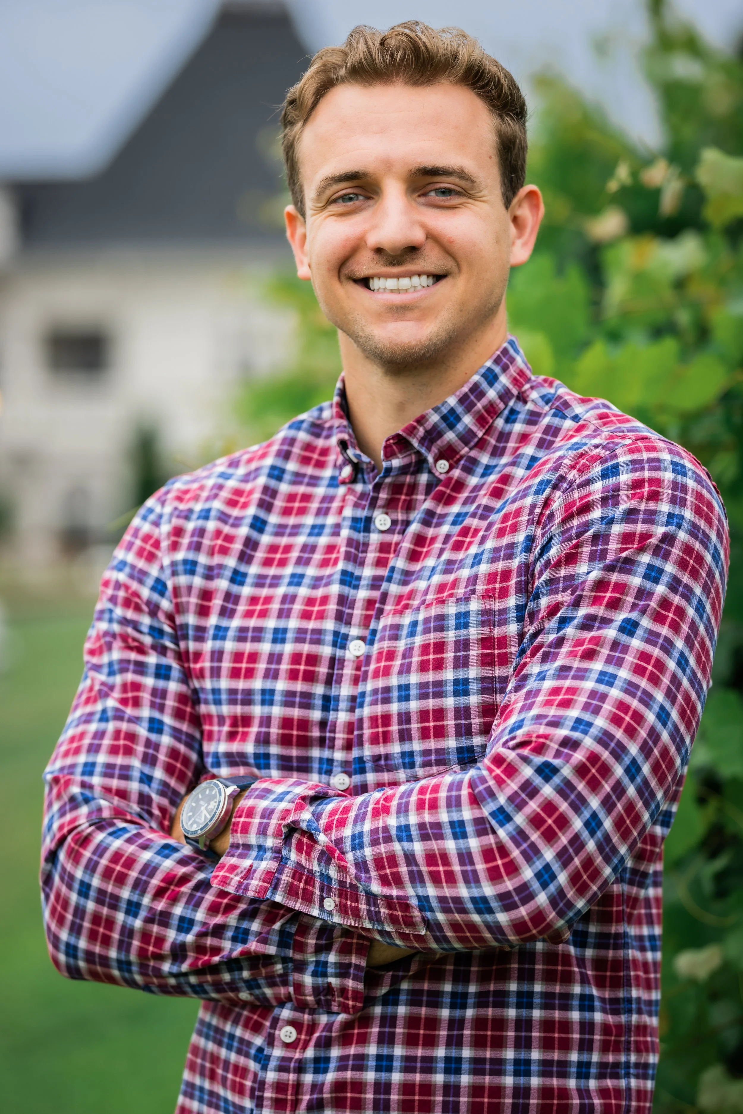 A man with short brown hair, smiling, standing outdoors with his arms crossed, wearing a red and blue plaid shirt and a watch on his left wrist. There is greenery and a house in the background.