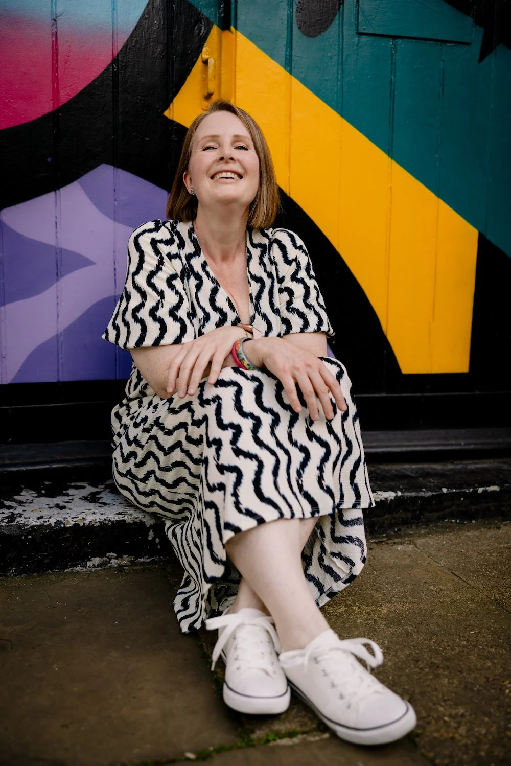A woman with shoulder-length brown hair, wearing a black and white wavy patterned dress and white sneakers, sitting on a concrete sidewalk with colorful graffiti on a wooden wall behind her, smiling at the camera.