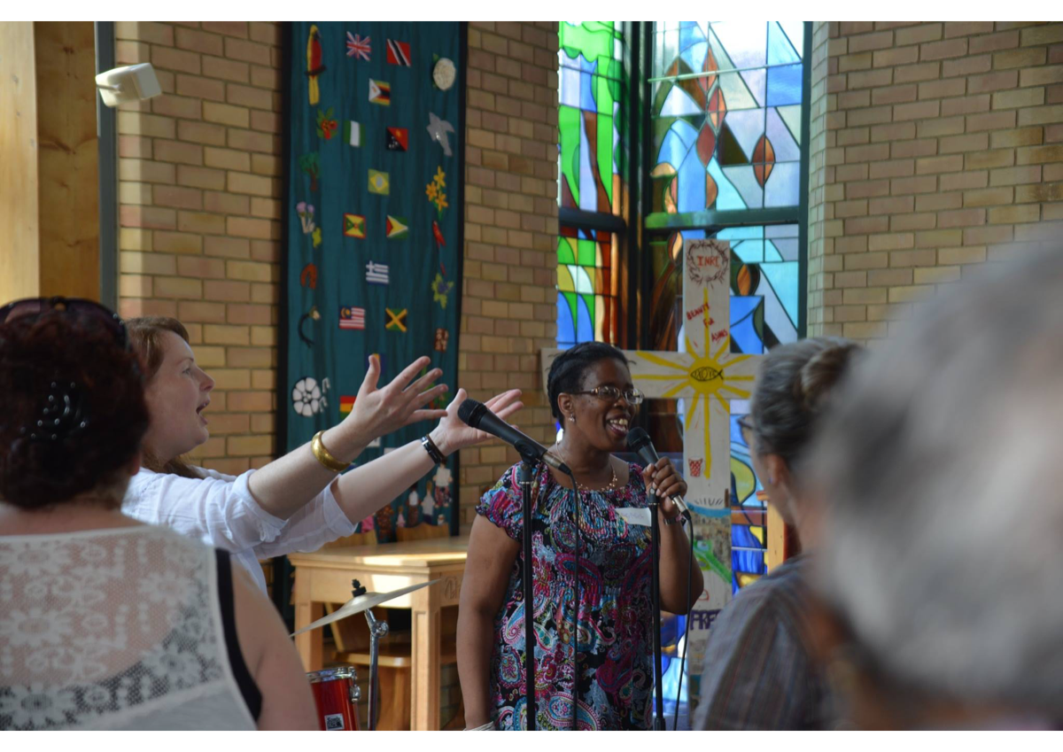A group of women gathered in a church for a presentation or speech. One woman is speaking into a microphone, smiling, while another woman gestures with her hands. The background features stained glass windows and a church banner depicting a cross with a dove.