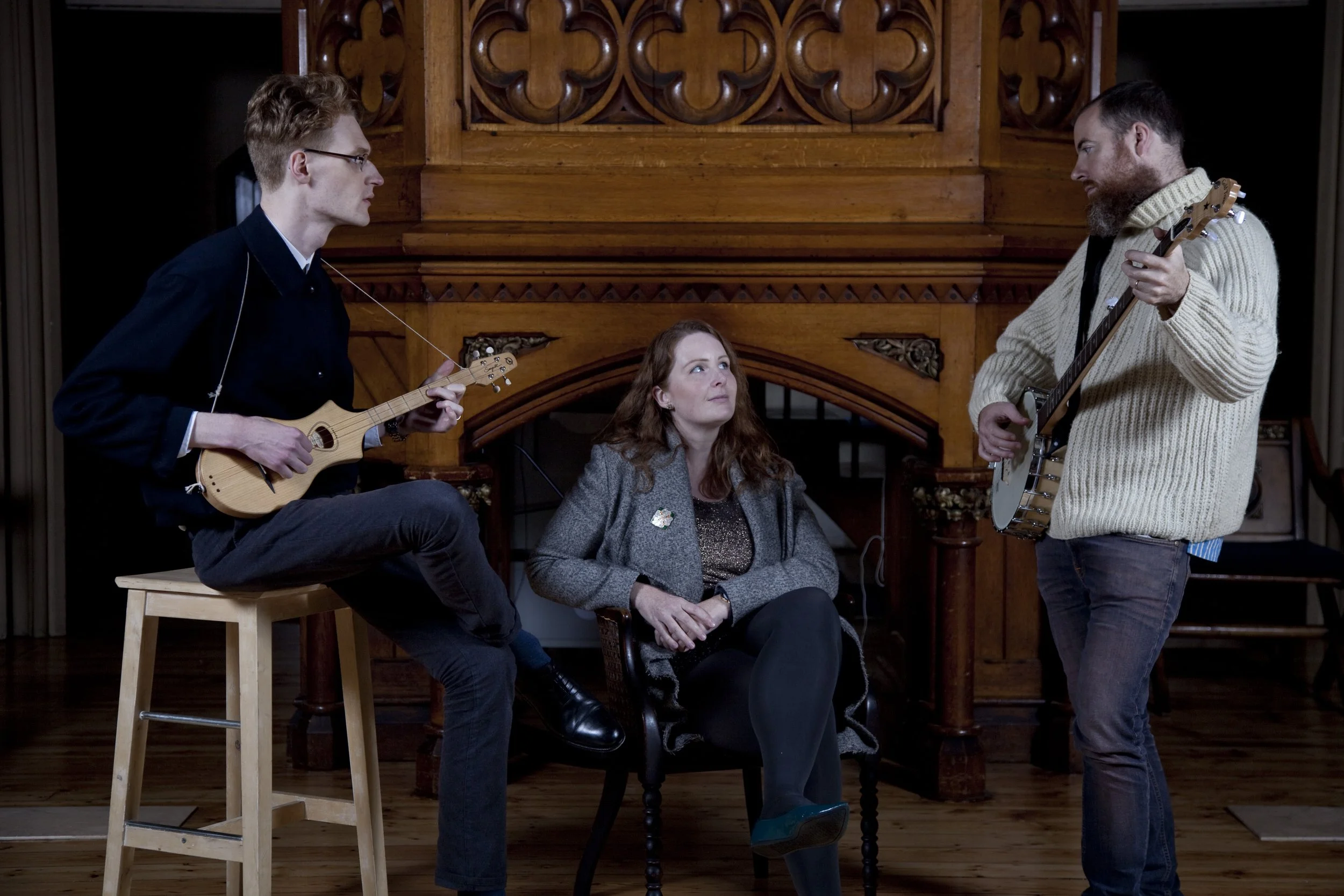 Three people in a room with wooden decor, two men playing guitars and a woman sitting between them, listening.