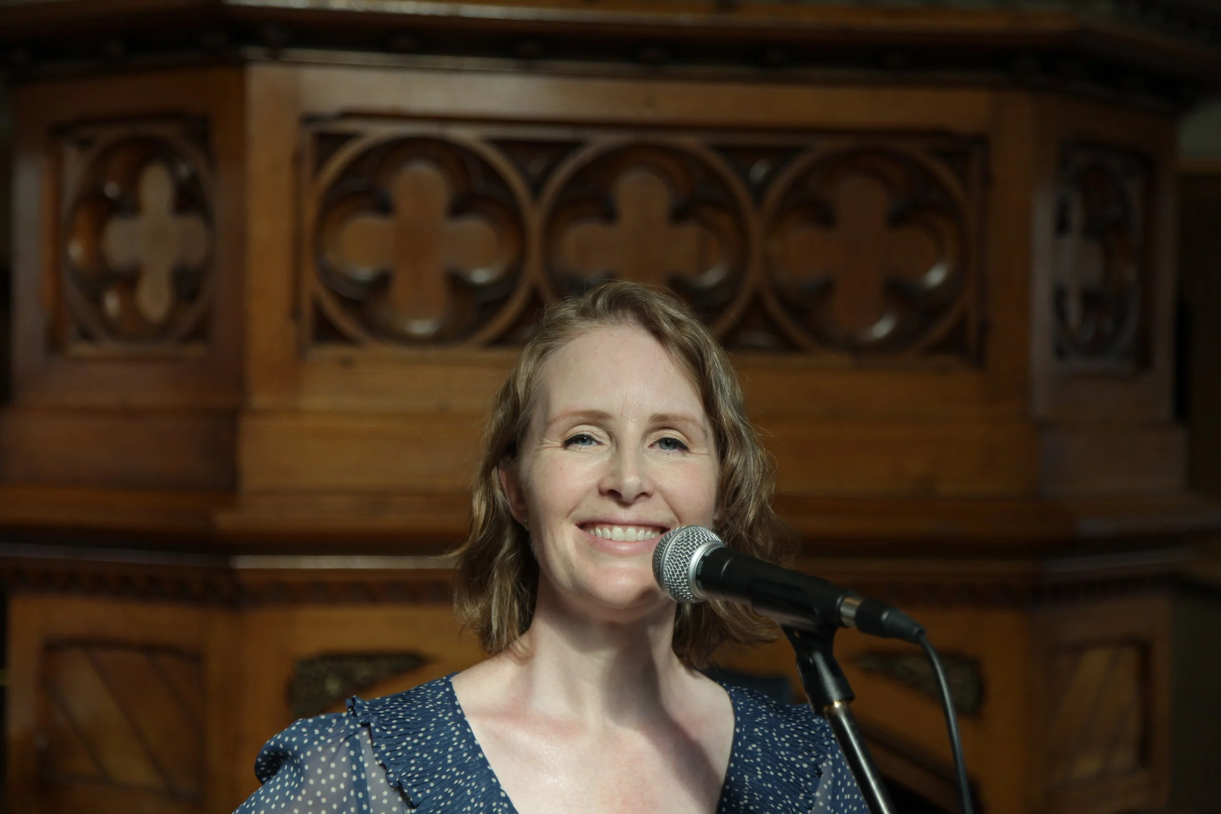 A woman with light brown curly hair smiling confidently while standing in front of a wooden decorative backdrop, speaking into a microphone.