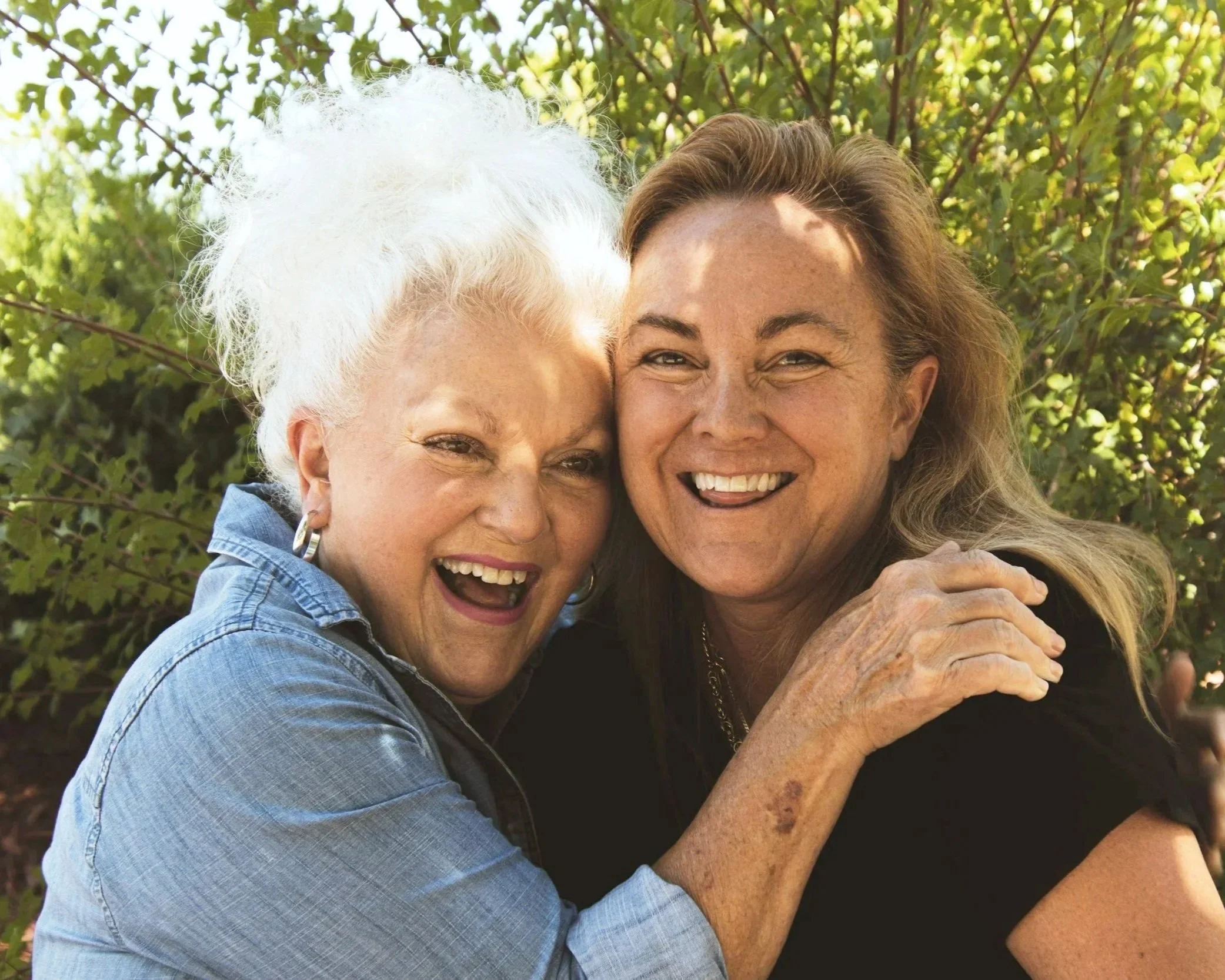 Two women hugging each other outdoors, smiling happily. One has white curly hair, and the other has long light brown hair. They are surrounded by green foliage.