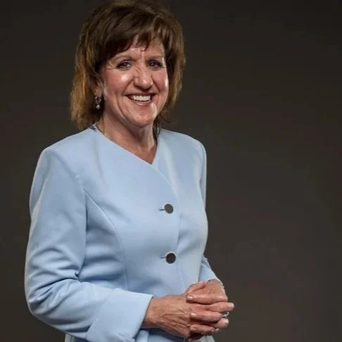 A middle-aged woman with short brown hair, smiling, wearing a light blue blazer, posing against a dark background.