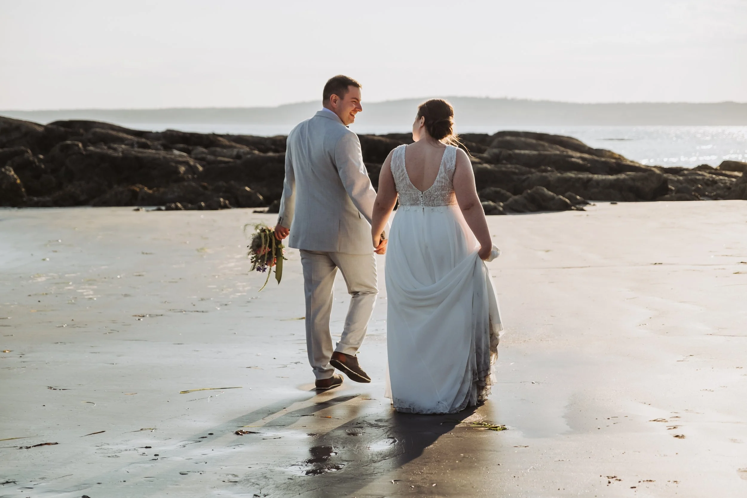 A newlywed couple walking hand in hand on a beach at sunset, with the bride in a white wedding dress and the groom in a light-colored suit, holding a bouquet, overlooking the ocean.