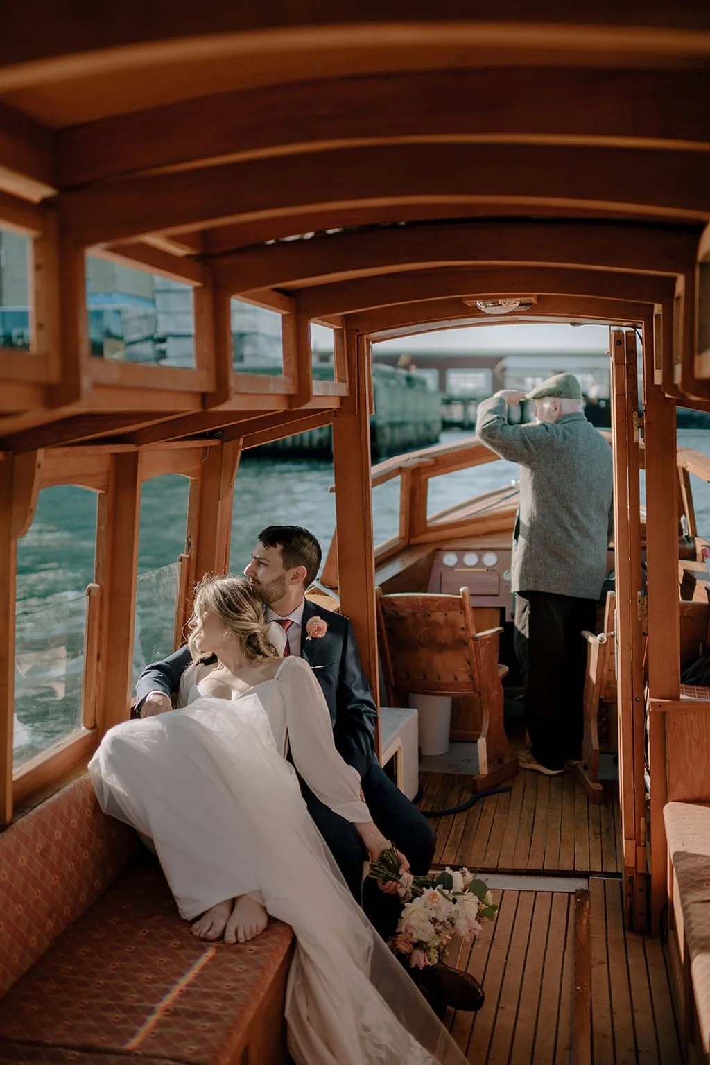 A couple in wedding attire sitting on a boat near a window, with the woman resting her head on the man's shoulder, and a man on the boat's bridge looking out through binoculars.