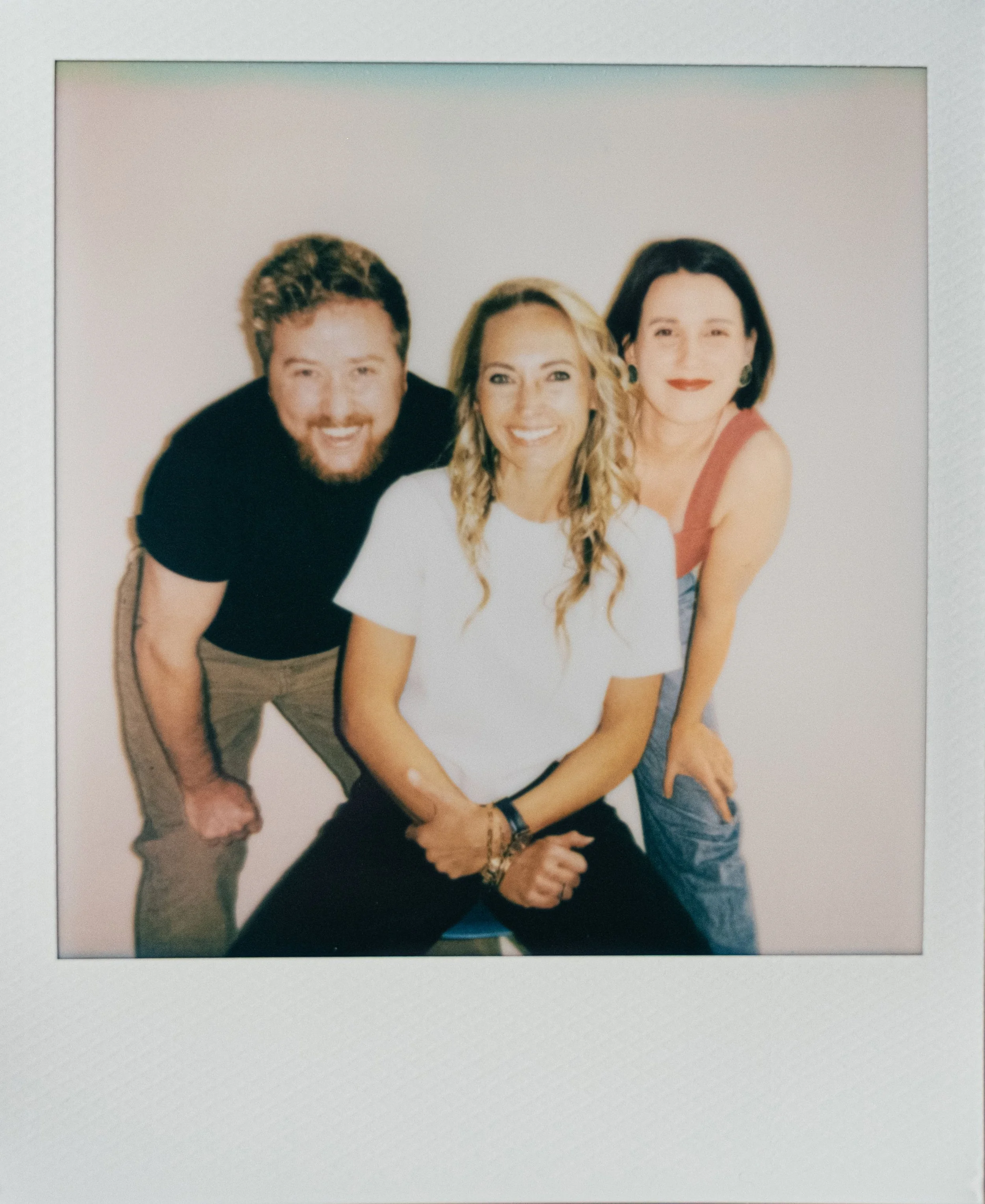 Three people posing together against a plain background, smiling and looking at the camera. They are a man with curly hair and beard, a woman with long curly blonde hair in the center, and a woman with short black hair wearing a red sleeveless top on the right.