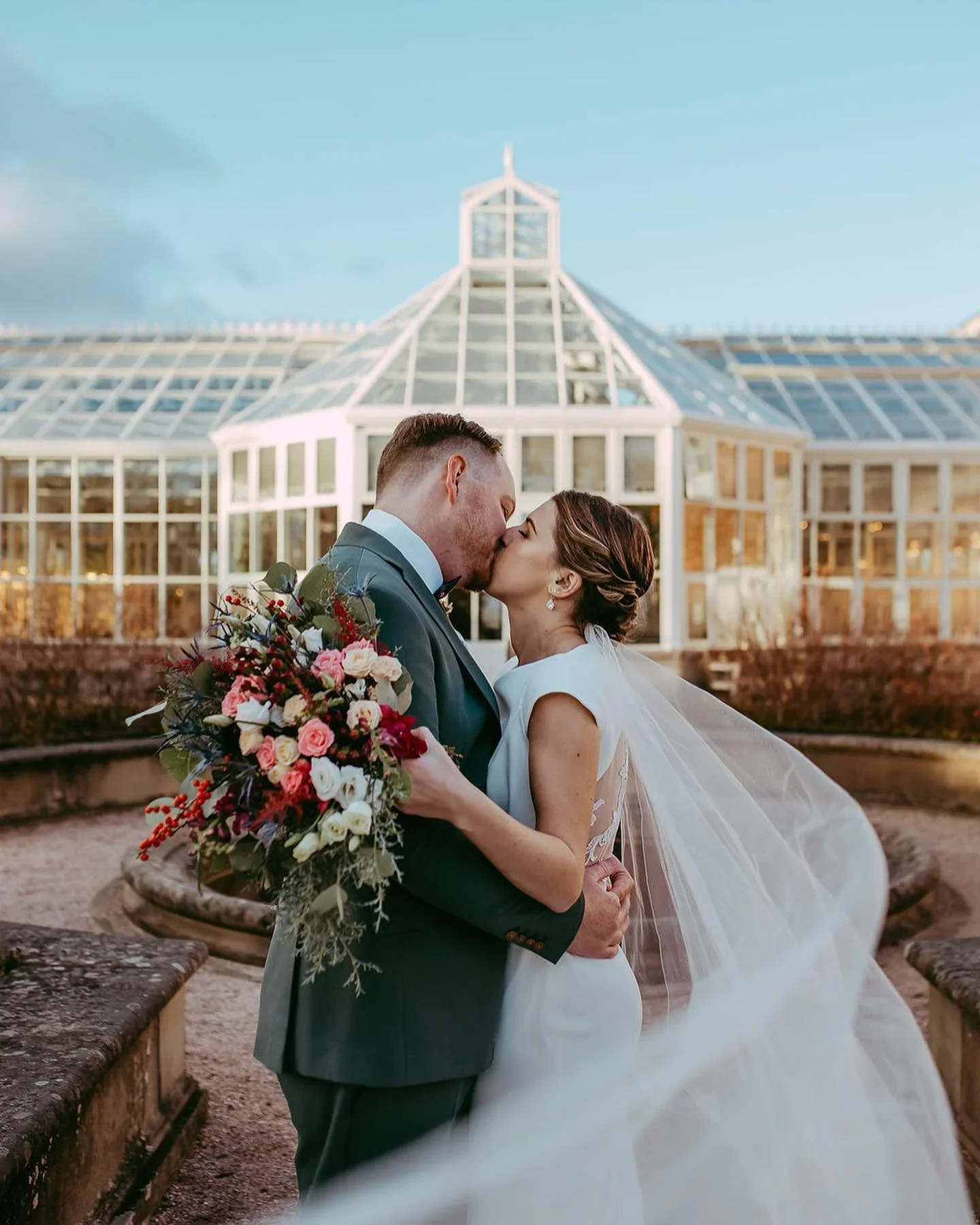 A newlywed couple sharing a kiss outdoors, with the bride holding a large bouquet of pink, white, and red flowers, standing in front of a glass conservatory under a blue sky.