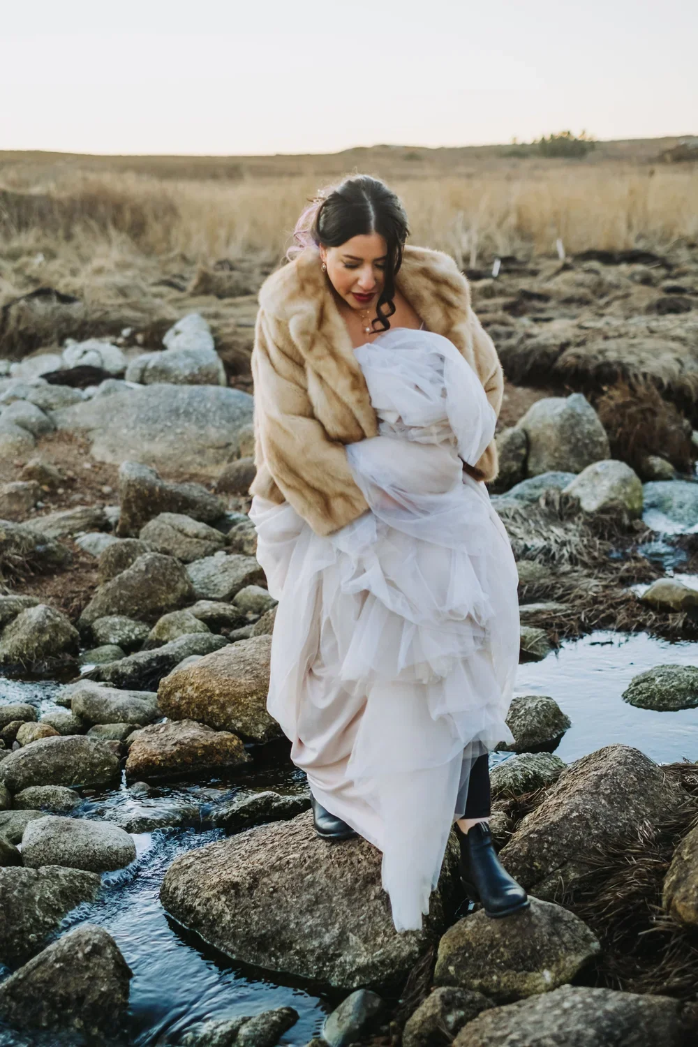 Woman in a wedding dress and fur coat stepping on rocks by a stream in an outdoor, rural setting.