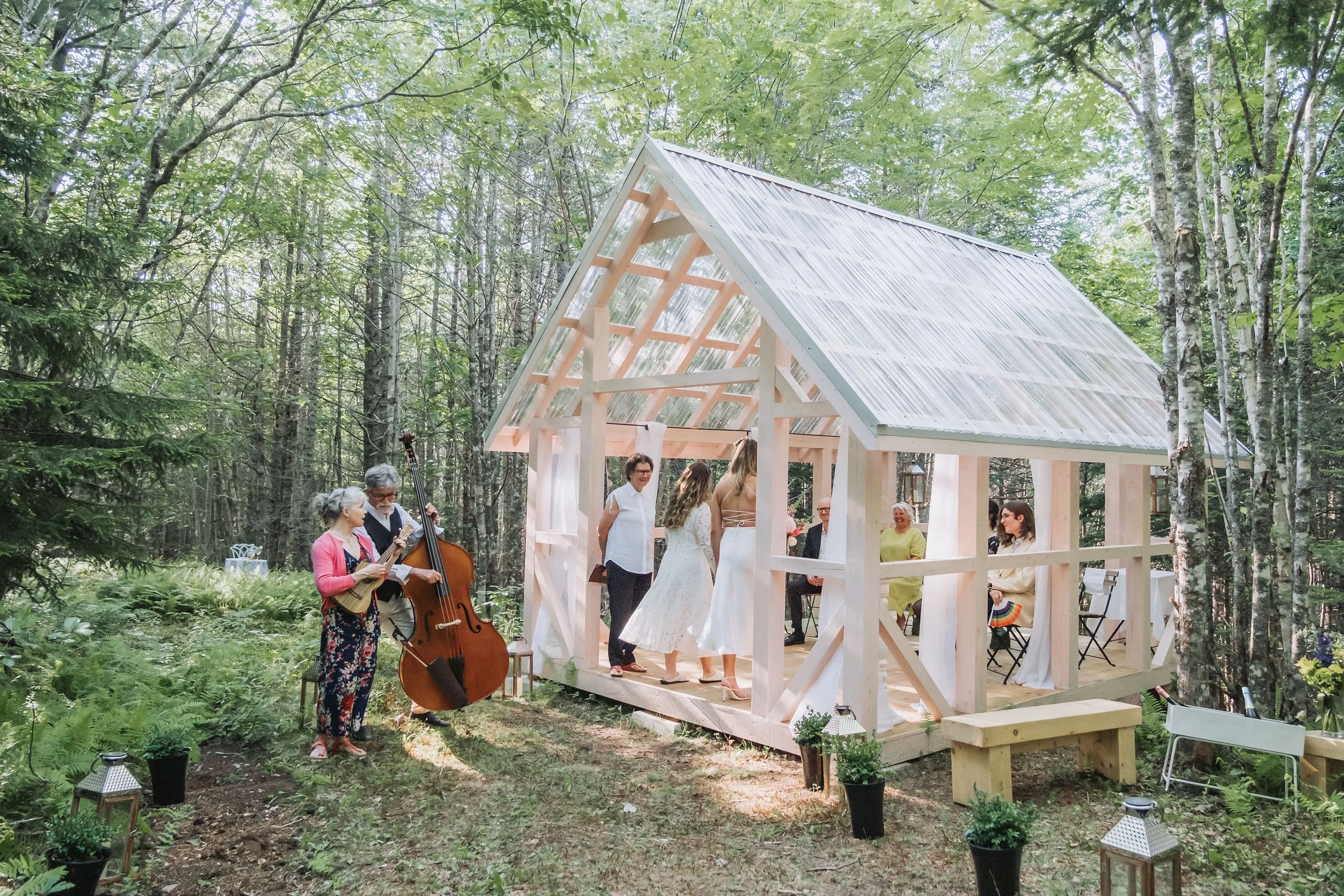 A wedding ceremony taking place outdoors in a forest setting. There are several people, including bridesmaids and family members, inside and outside a small wooden chapel under construction with a translucent roof. Musicians playing guitar and double bass are outside near the chapel. Decorations include string lights and potted plants along the ground.