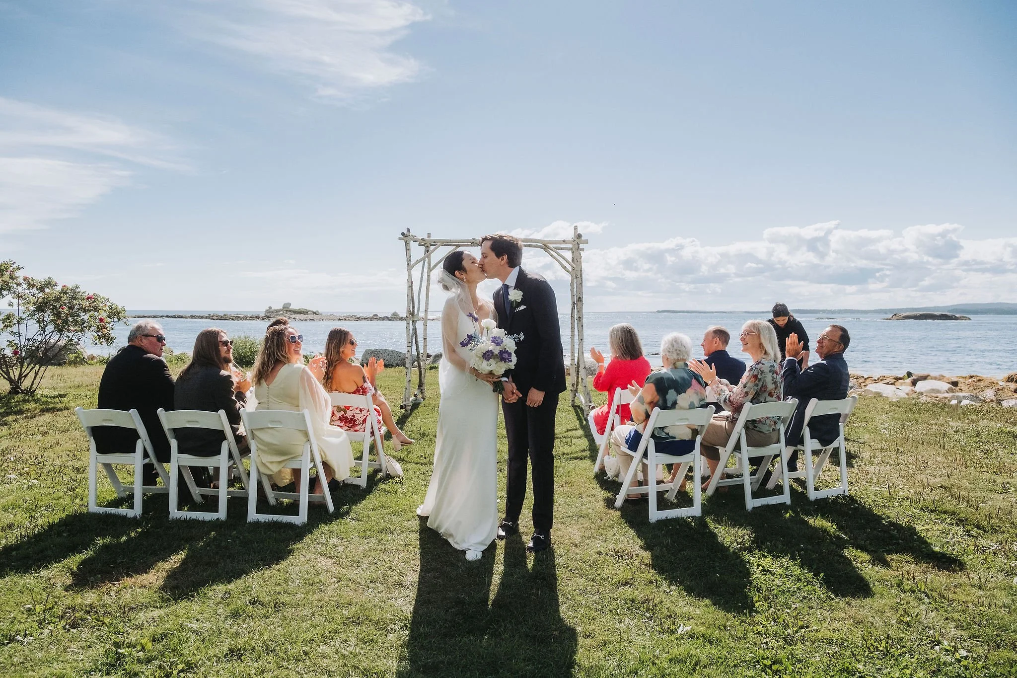 A couple gets married outdoors by the water, sharing a kiss under a wooden arch, surrounded by friends and family seated on white chairs, with a scenic ocean view and blue sky in the background.