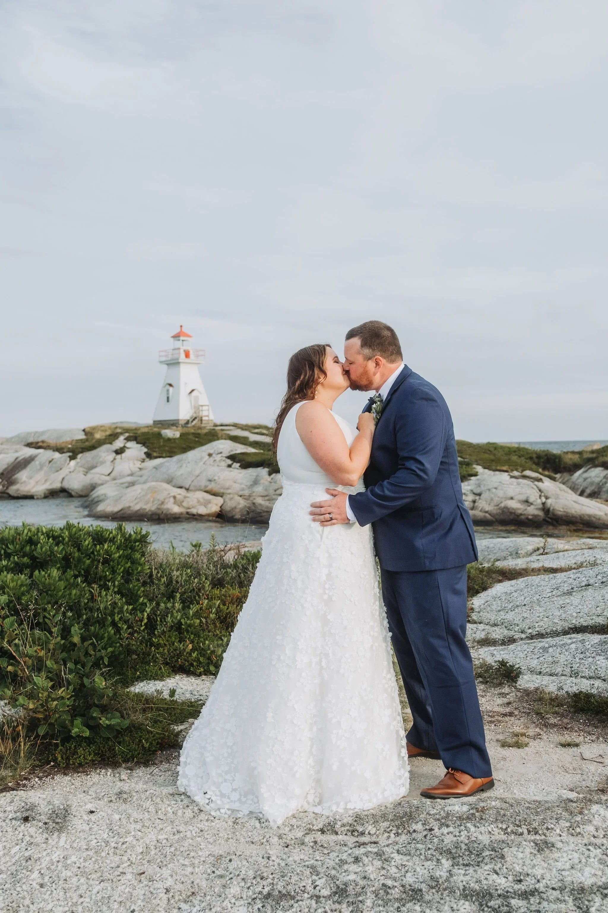 A couple in wedding attire share a kiss by the ocean with a lighthouse in the background.