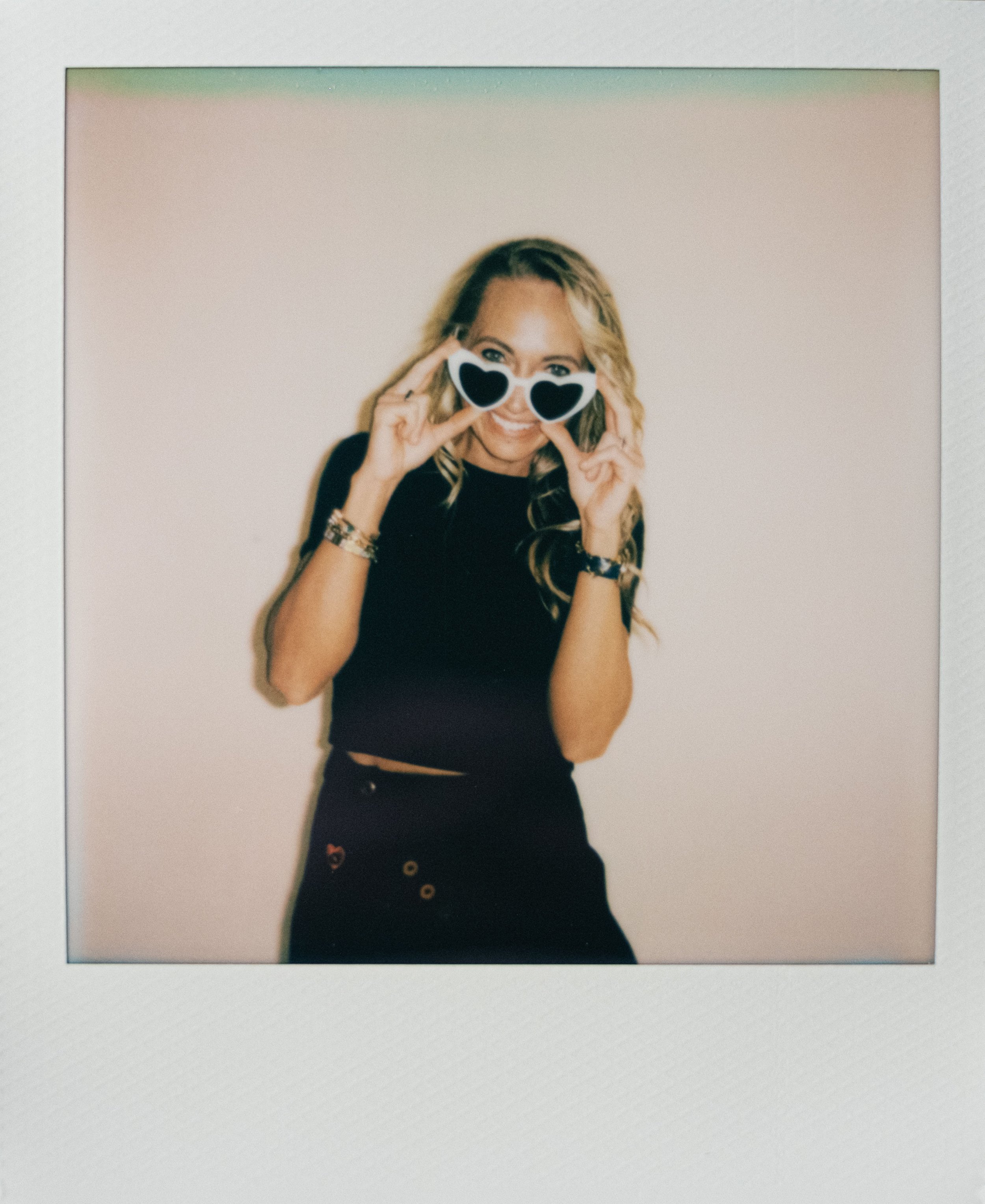 Woman with blonde hair wearing heart-shaped sunglasses, black top, and black pants smiling and posing against a plain background.