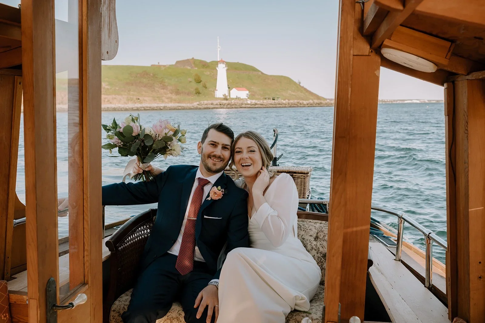 A newlywed couple on a boat, smiling and holding a bouquet of flowers, with water and a lighthouse on a hill in the background.