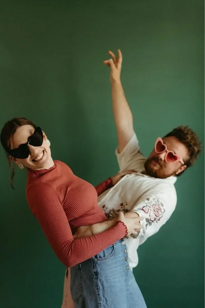A woman and a man with sunglasses and playful outfits dancing together against a green background.