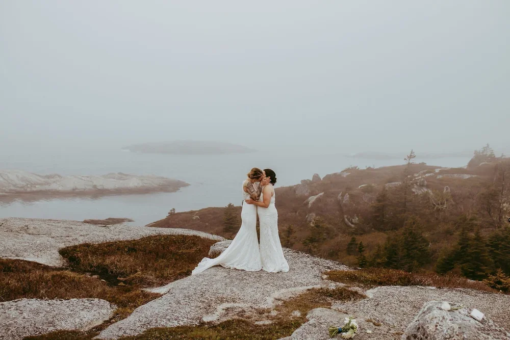 Two brides in wedding dresses embracing and kissing on a rocky mountain with water and fog in the background.