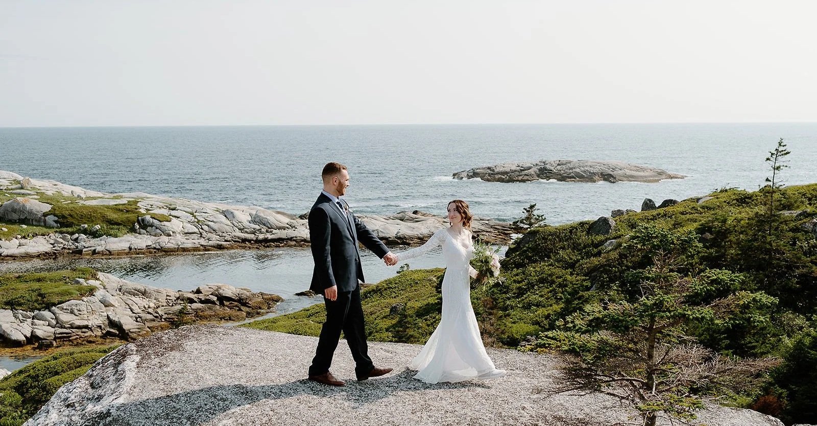 A bride and groom holding hands on a rocky coastal landscape with the ocean in the background and the bride holding a bouquet.