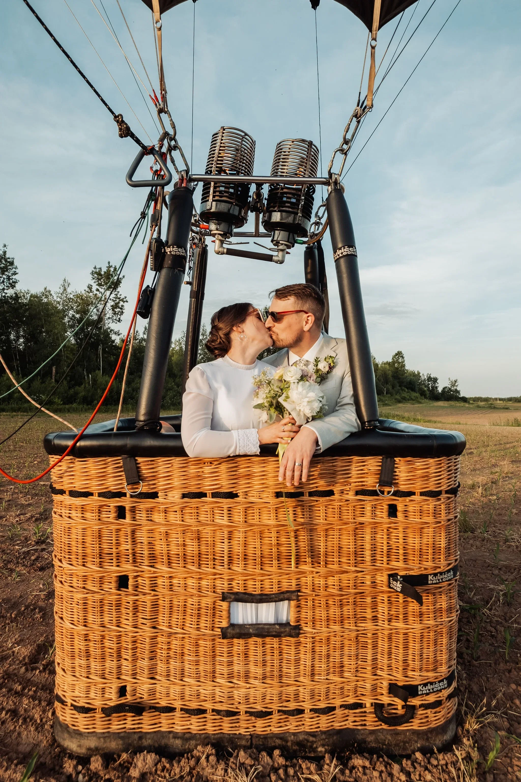 A couple in wedding attire kissing inside a hot air balloon basket, holding a bouquet of flowers, with the balloon's burner above them and a rural landscape in the background.