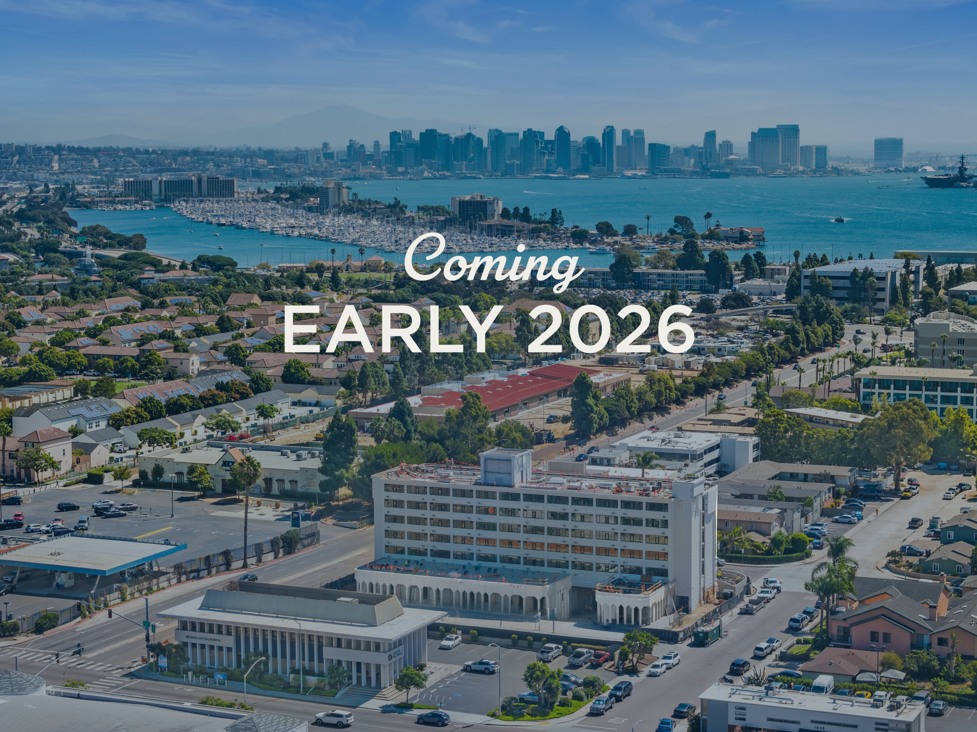 A cityscape with a skyline of high-rise buildings across a body of water, surrounded by a residential area with houses and greenery, advertising an upcoming project in early 2026.