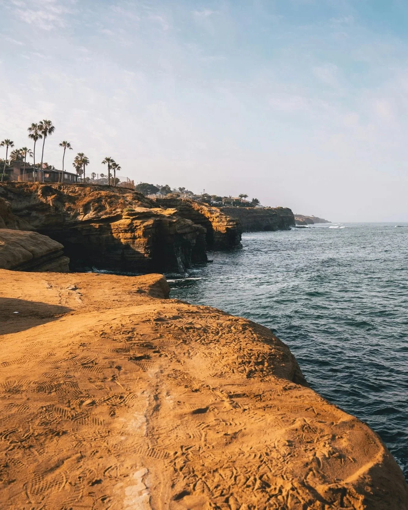 Cliffs with sandy ledge, ocean waves, and houses with palm trees on top, under a partly cloudy sky.