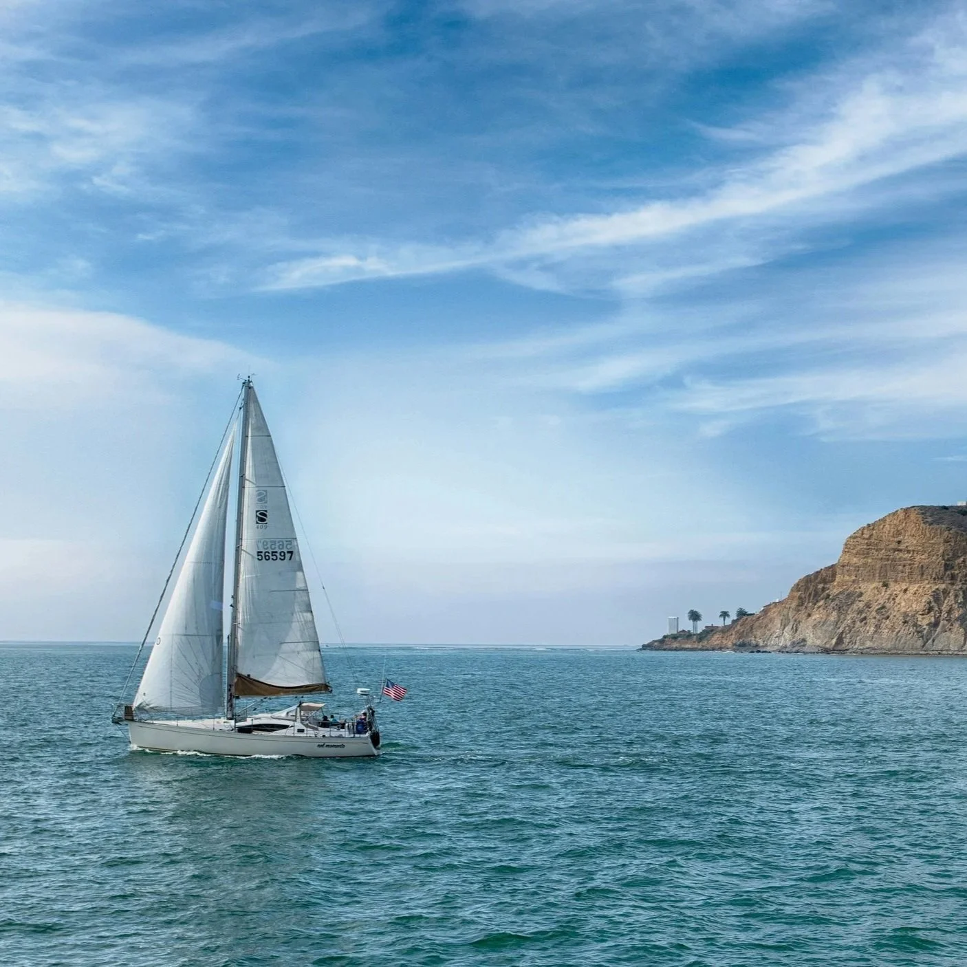 A sailboat with white sails and an American flag sailing near rocky cliffs on a calm ocean under a blue sky with scattered clouds.
