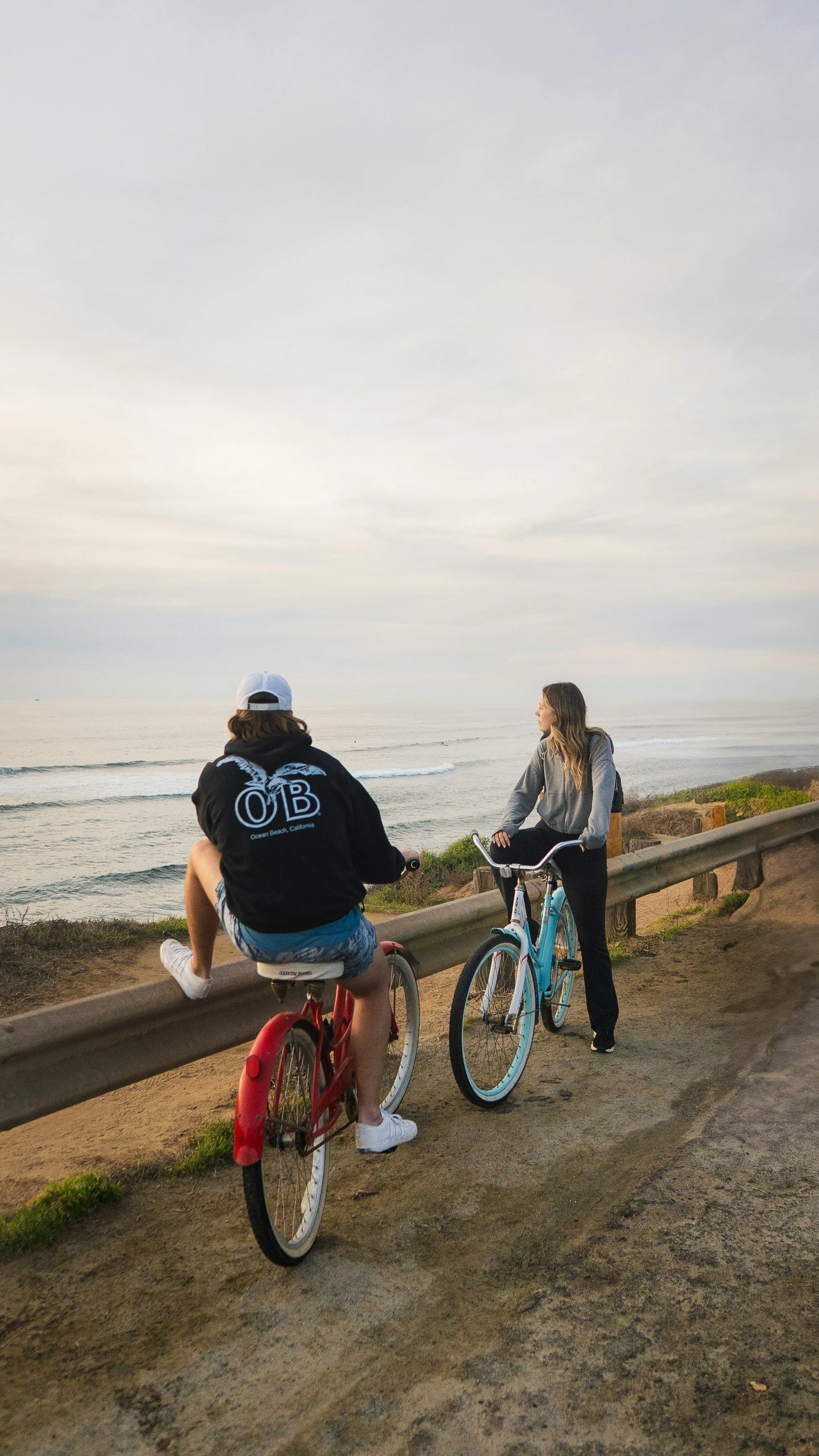 Two women with bicycles at a scenic seaside overlook, one sitting on a red bike and the other standing with a blue bike, during sunset.