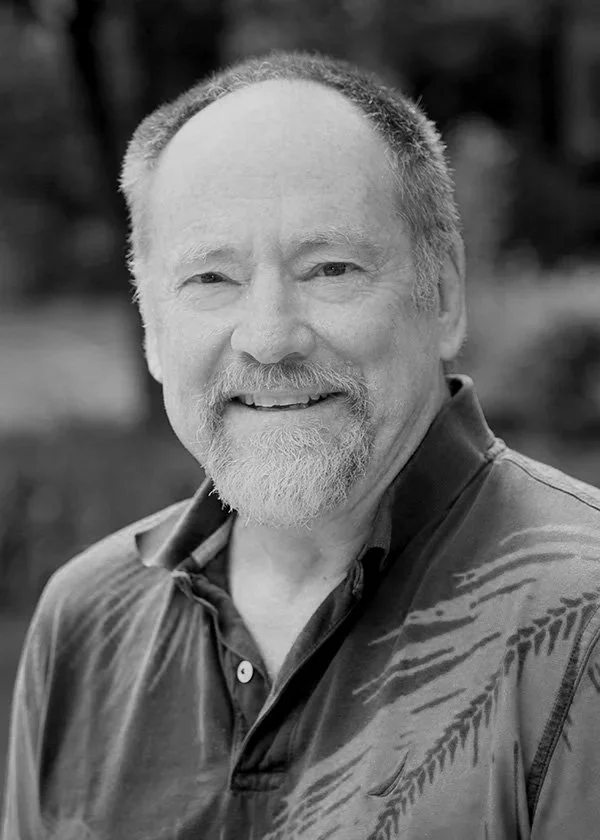 Black and white photograph of a middle-aged man with a beard and mustache, smiling and wearing a collared shirt. The background is blurred, suggesting an outdoor setting.