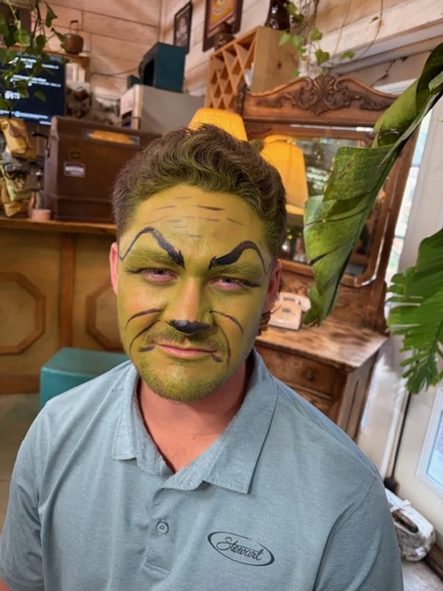 Man with face painted as a tiger, wearing a light blue polo shirt, indoors with wooden decor, plants, and warm lighting.