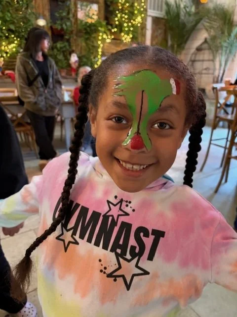 Young girl with a big smile, face painted with a Grinch hand on forehead, hangingan ornament on her nose.